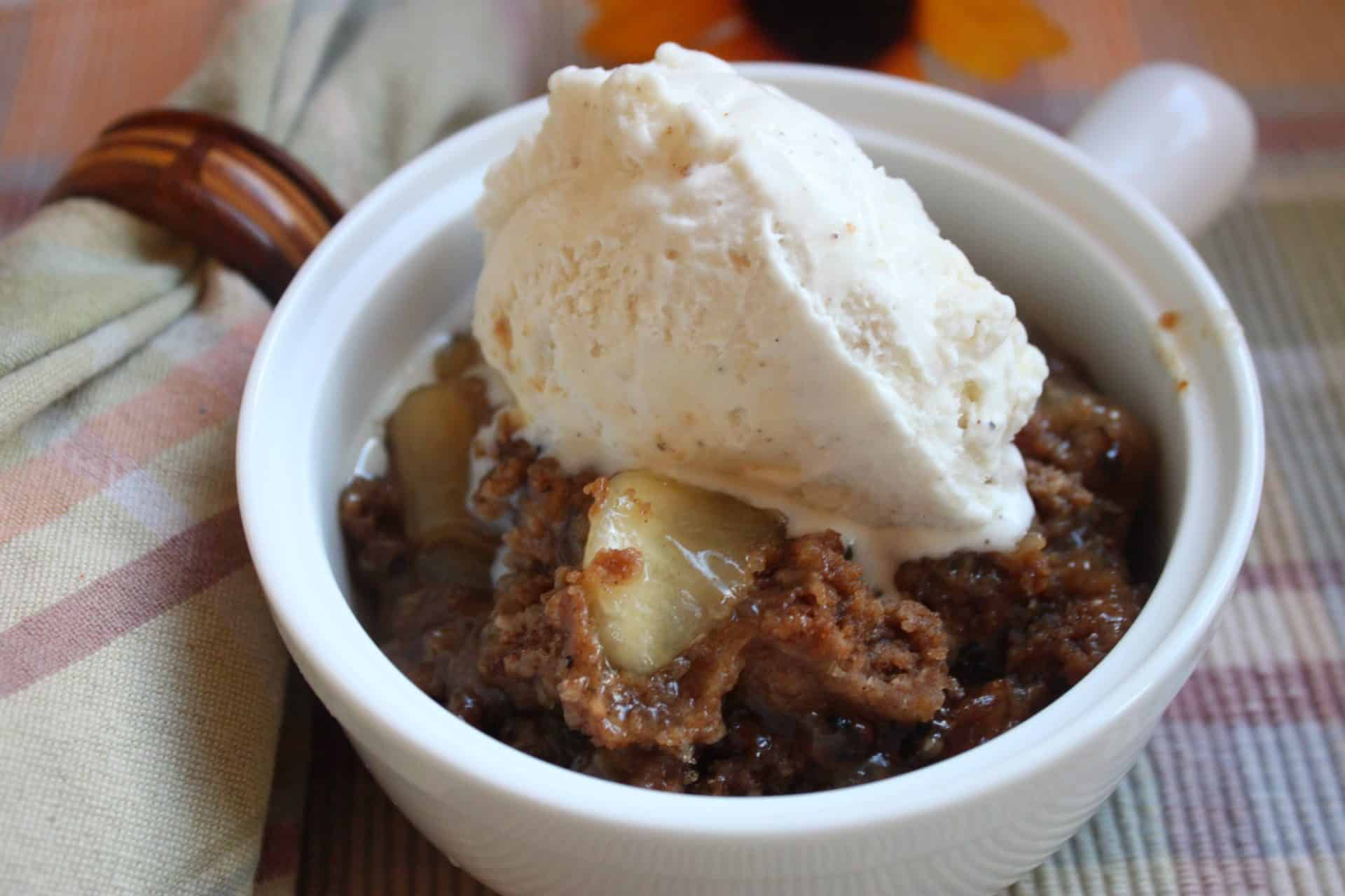 A full serving of 3 Step Crockpot Apple Spice Cake in a white bowl with a scoop of vanilla ice cream, perfectly melted over the warm dessert.