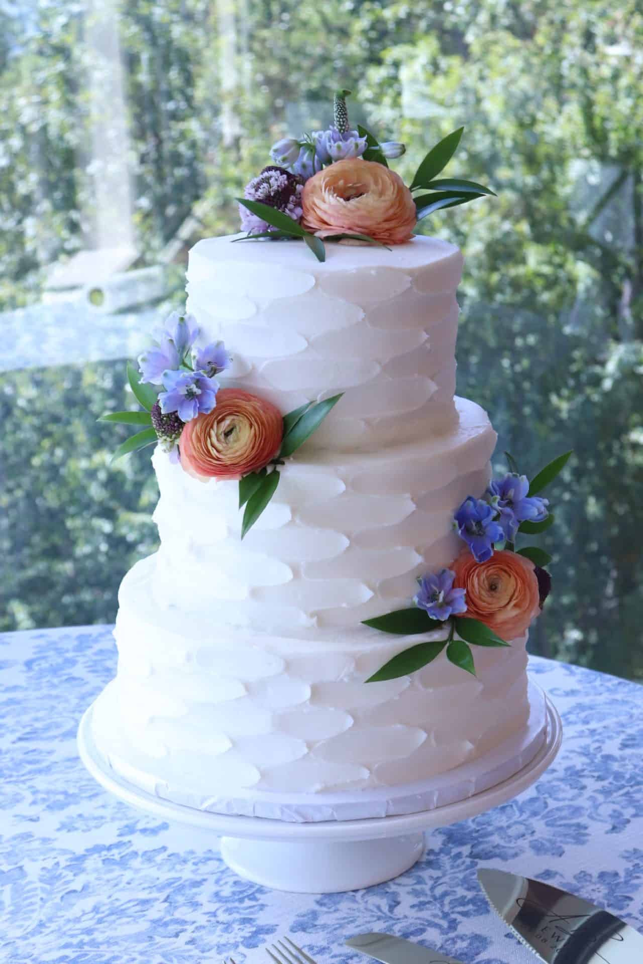 Cake with palette knife-textured white frosting, adorned with peachy-pink ranunculus flowers, vibrant blue blossoms, and greenery accents.