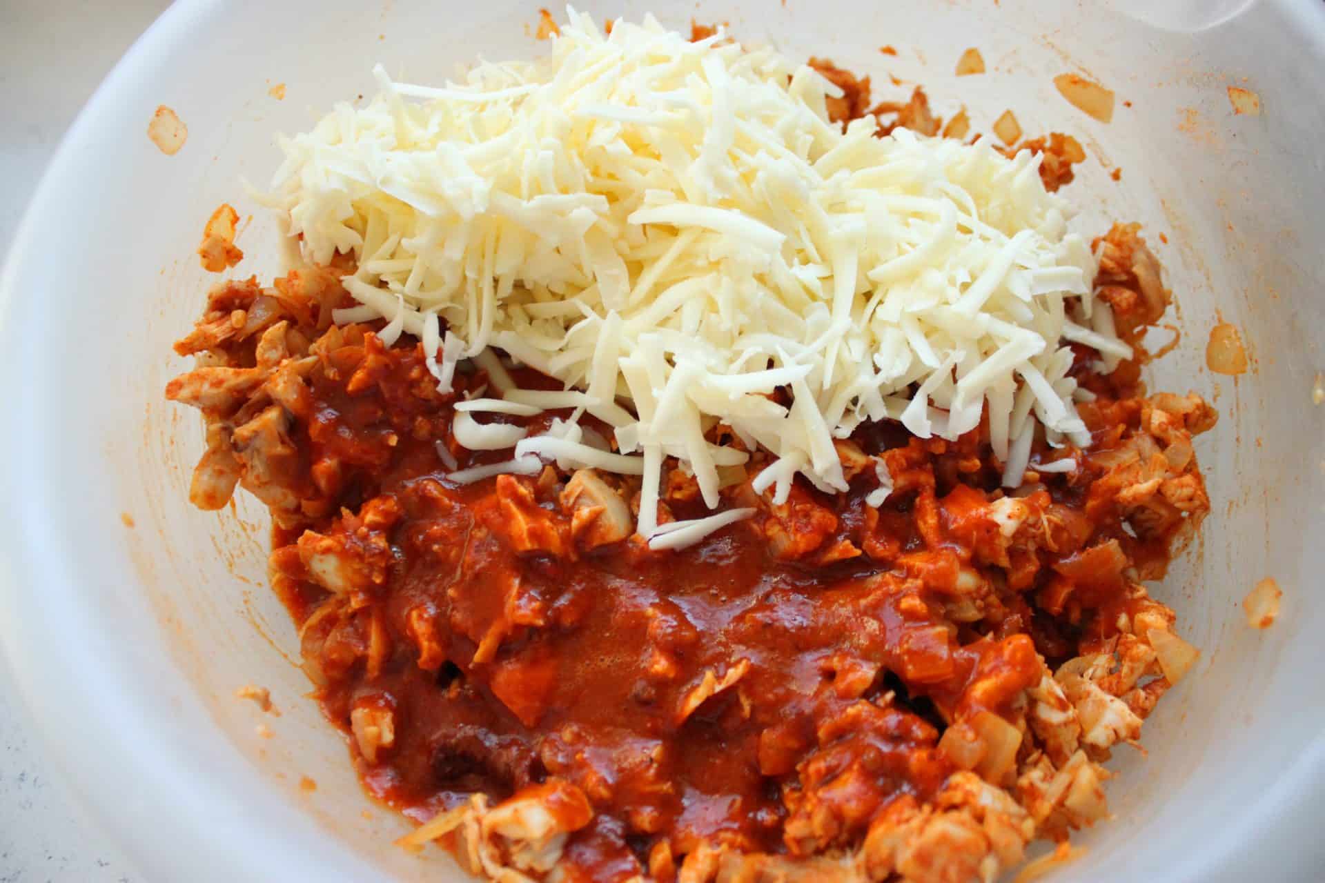 Tortillas filled with spicy chicken mixture, topped with red sauce and shredded cheese, ready for the oven.