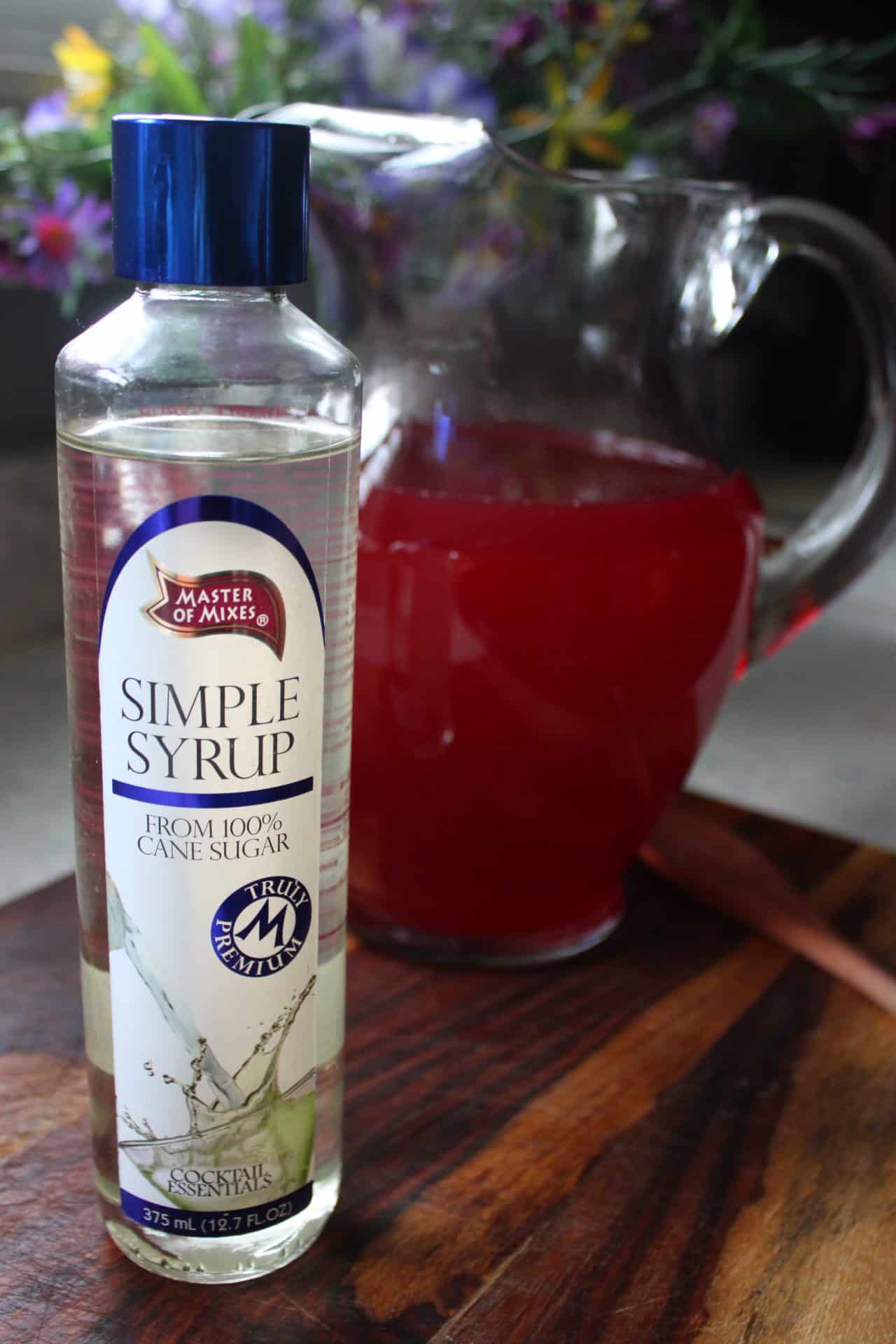 Bottle of simple syrup next to a pitcher of bright pink Starbucks Iced Passion Tea Lemonade, ready to sweeten.