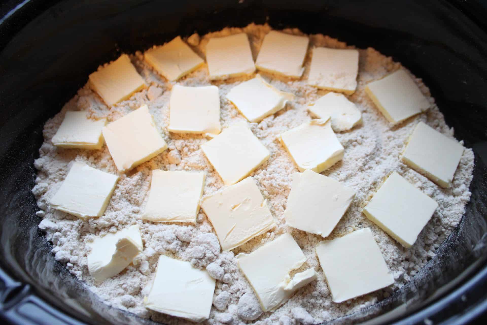 Chunks of cold butter arranged evenly over a layer of dry cake mix inside a Crockpot during prep for 3 Step Crockpot Apple Spice Cake.