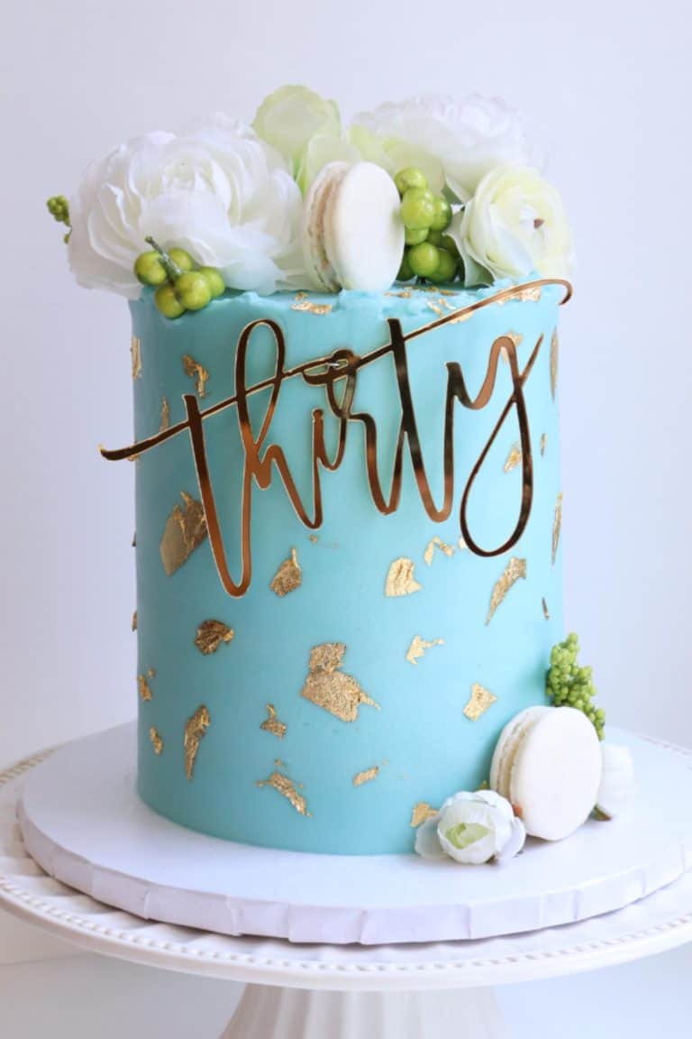 Sophisticated cake with blue fondant accented by gold leaf, topped with white flowers, macarons, and a golden "thirty" topper.