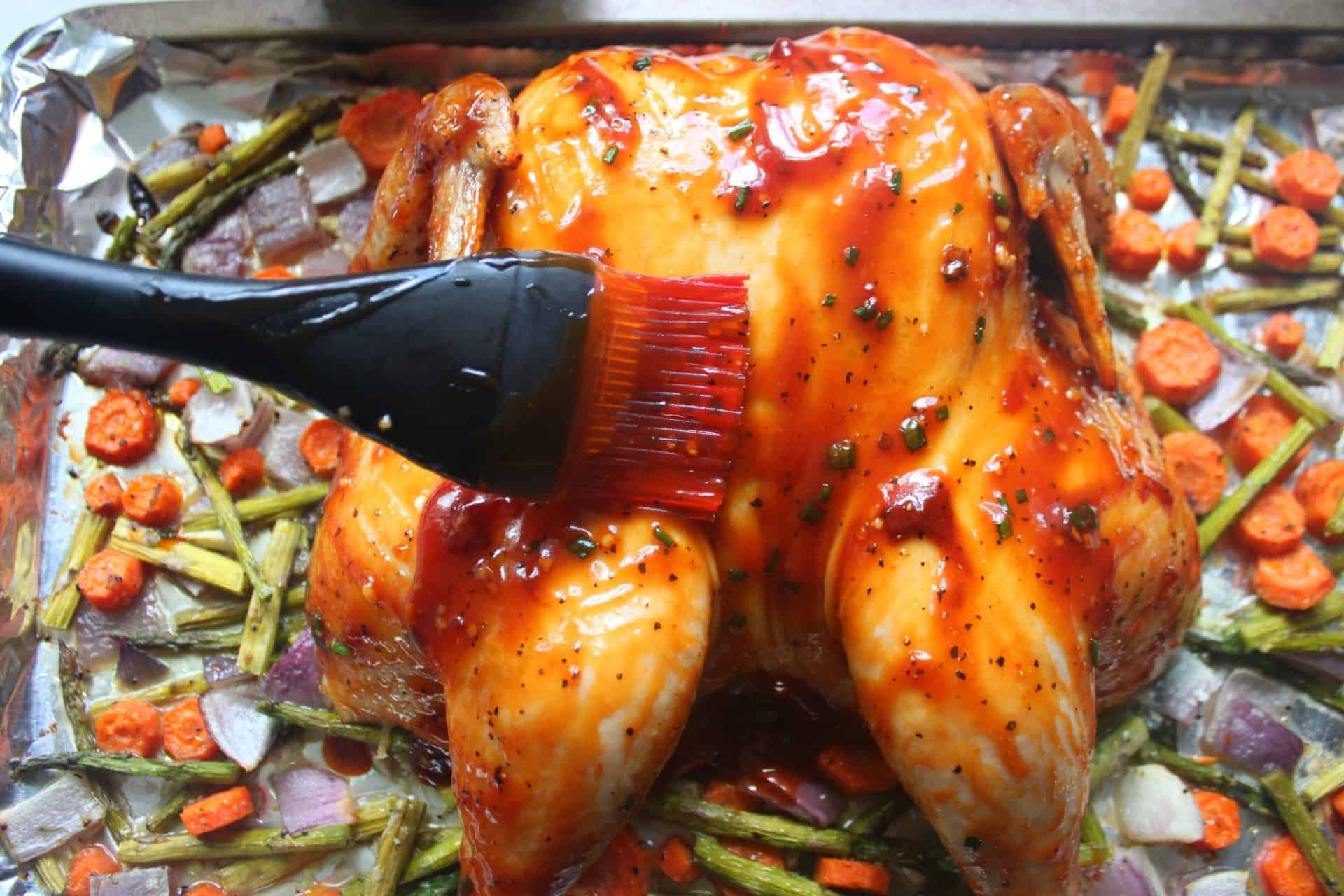 Basting brush applying thick BBQ sauce over a whole spatchcock chicken just before roasting with vegetables.