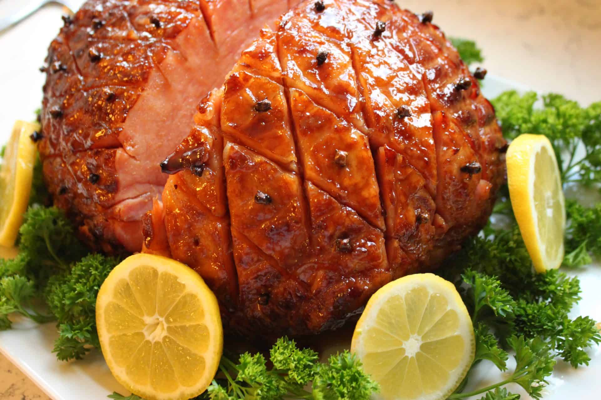 Whole baked ham with a shiny Coca-Cola glaze and diamond-cut pattern, surrounded by lemon slices and parsley for Easter dinner.