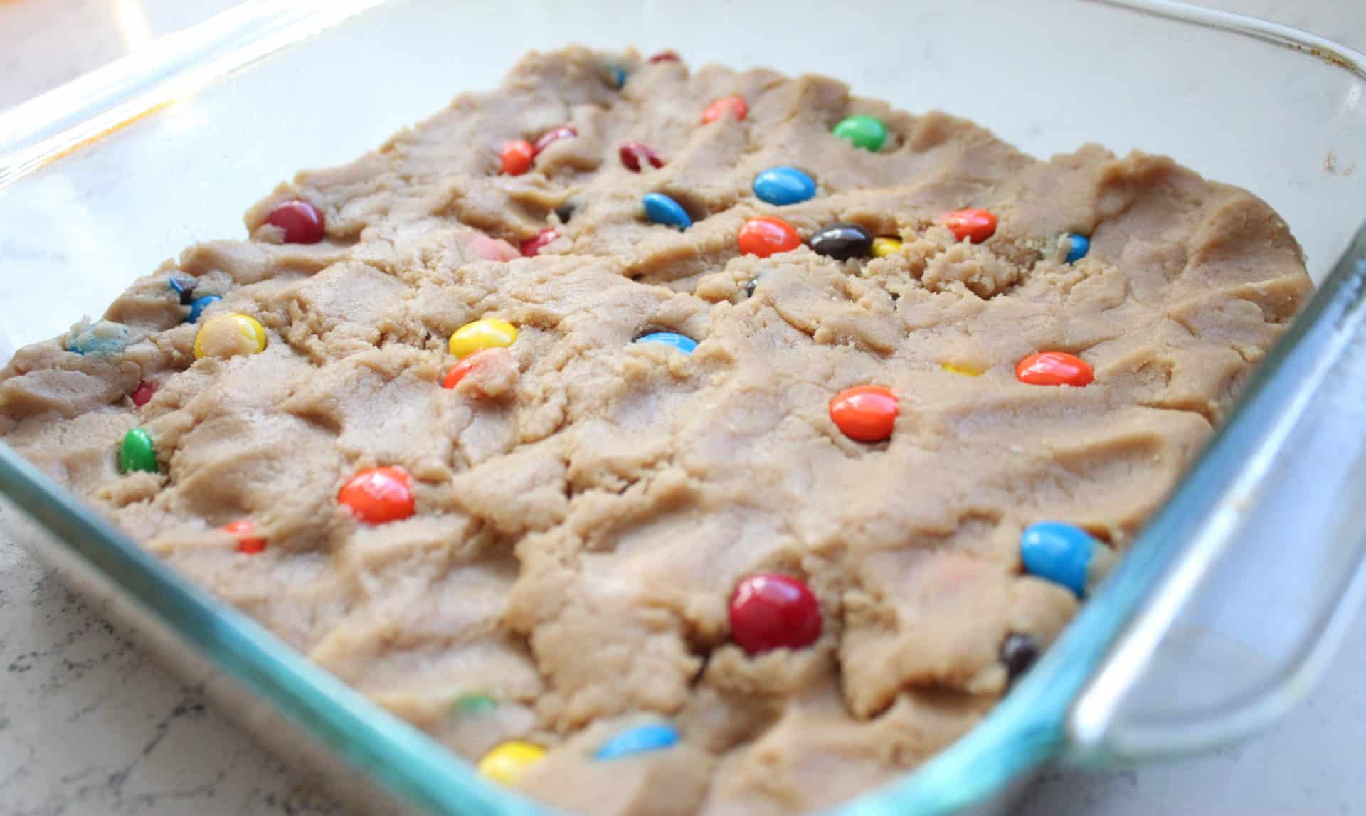 Irresistible M&M Peanut Butter Bars: 5 Simple Ingredients 9 M&M Peanut Butter Bar Dough Spread out in a Clear Glass Dish