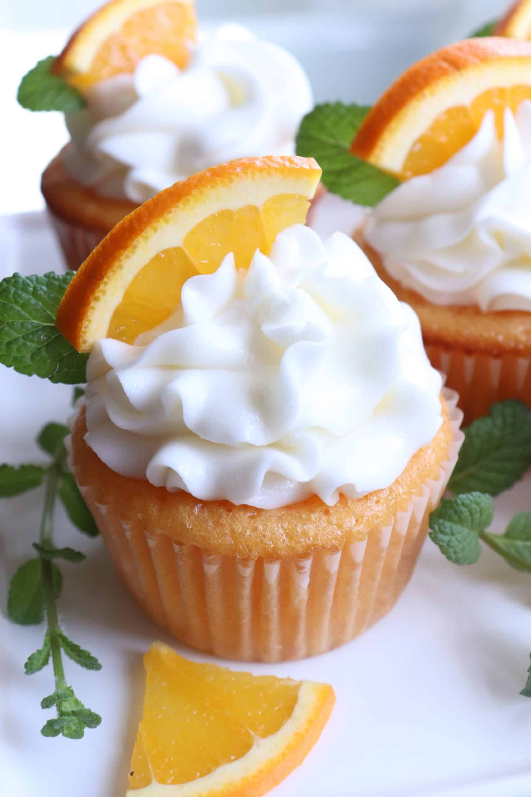 A Dreamsicle cupcake with white buttercream swirl, topped with a fresh orange slice and mint sprig, surrounded by extra mint and orange pieces.