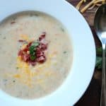 A bowl of Potato Cheese Soup topped with crispy bacon, shredded cheese, and fresh herbs, served hot for a delicious meal.