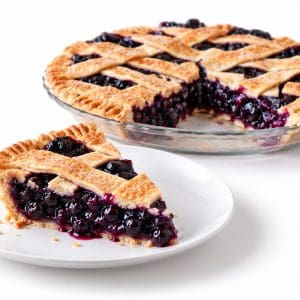 Slice of blueberry pie on a white plate with juicy berry filling and the remaining lattice pie behind it.