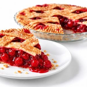 Slice of cherry lattice pie on a white plate with glossy red filling and crumbs, with the remaining pie softly blurred in a glass dish behind it.