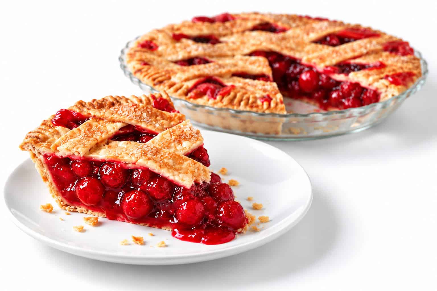 Slice of cherry lattice pie on a white plate with glossy red filling and crumbs, with the remaining pie softly blurred in a glass dish behind it.