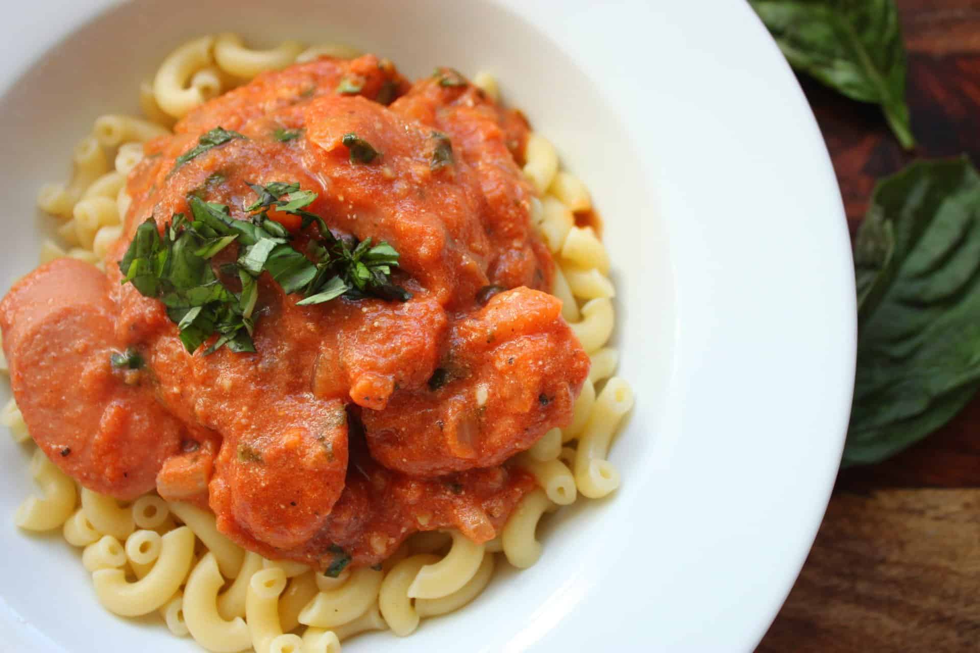 Close-up of a white plate filled with kielbasa basil pasta, showcasing tender sausage slices and fresh herbs on top.