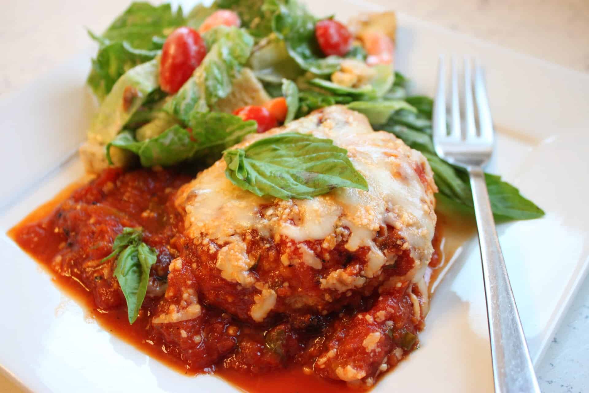 Close-up of Chicken Parmesan topped with cheese and basil, served beside a crisp green salad with tomatoes on a white plate.