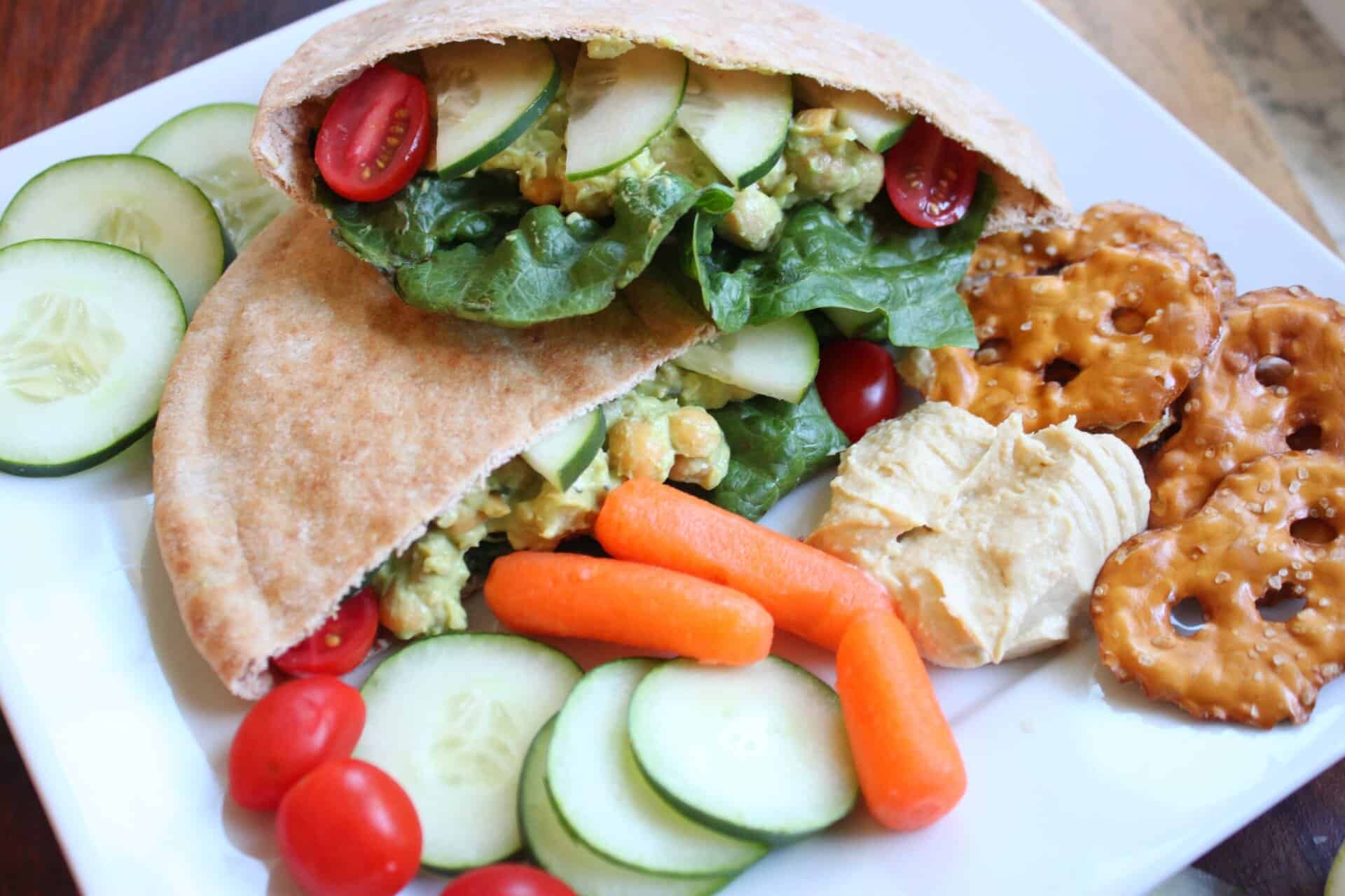 Whole wheat pita pocket stuffed with avocado chickpea filling, lettuce, cucumbers, and tomatoes on a plate with carrots and hummus.