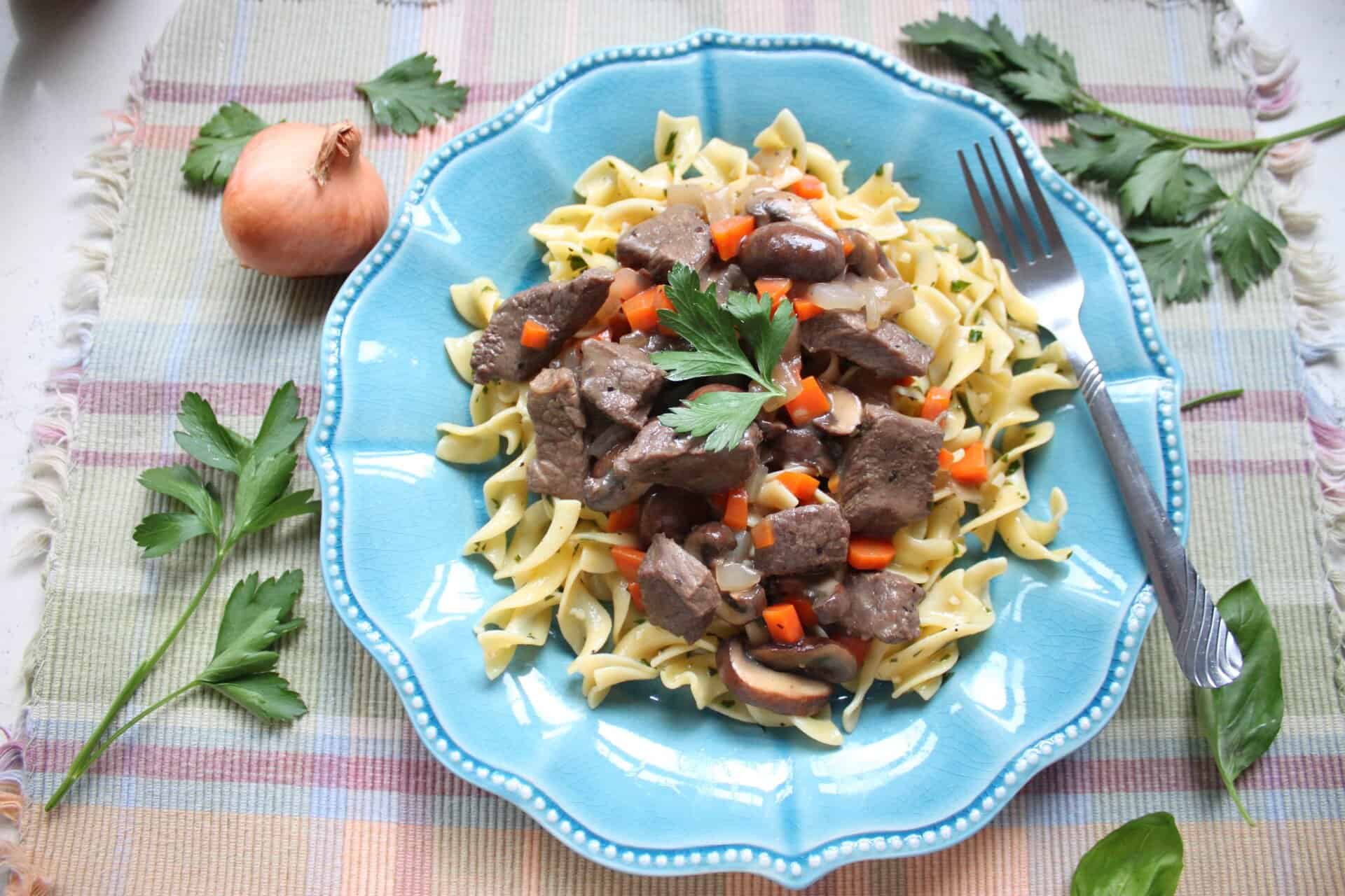 Plate of beef stroganoff with buttered noodles, garnished with chopped parsley and served on a blue scalloped plate with a silver fork.