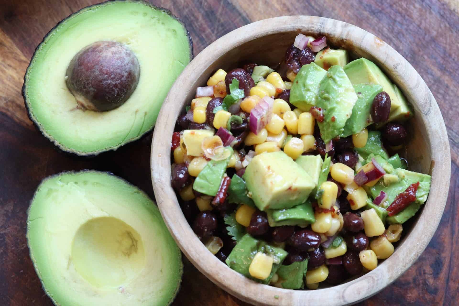 Black bean and corn salad with chipotle dressing served in a wooden bowl with fresh diced avocado and whole avocados in the background.