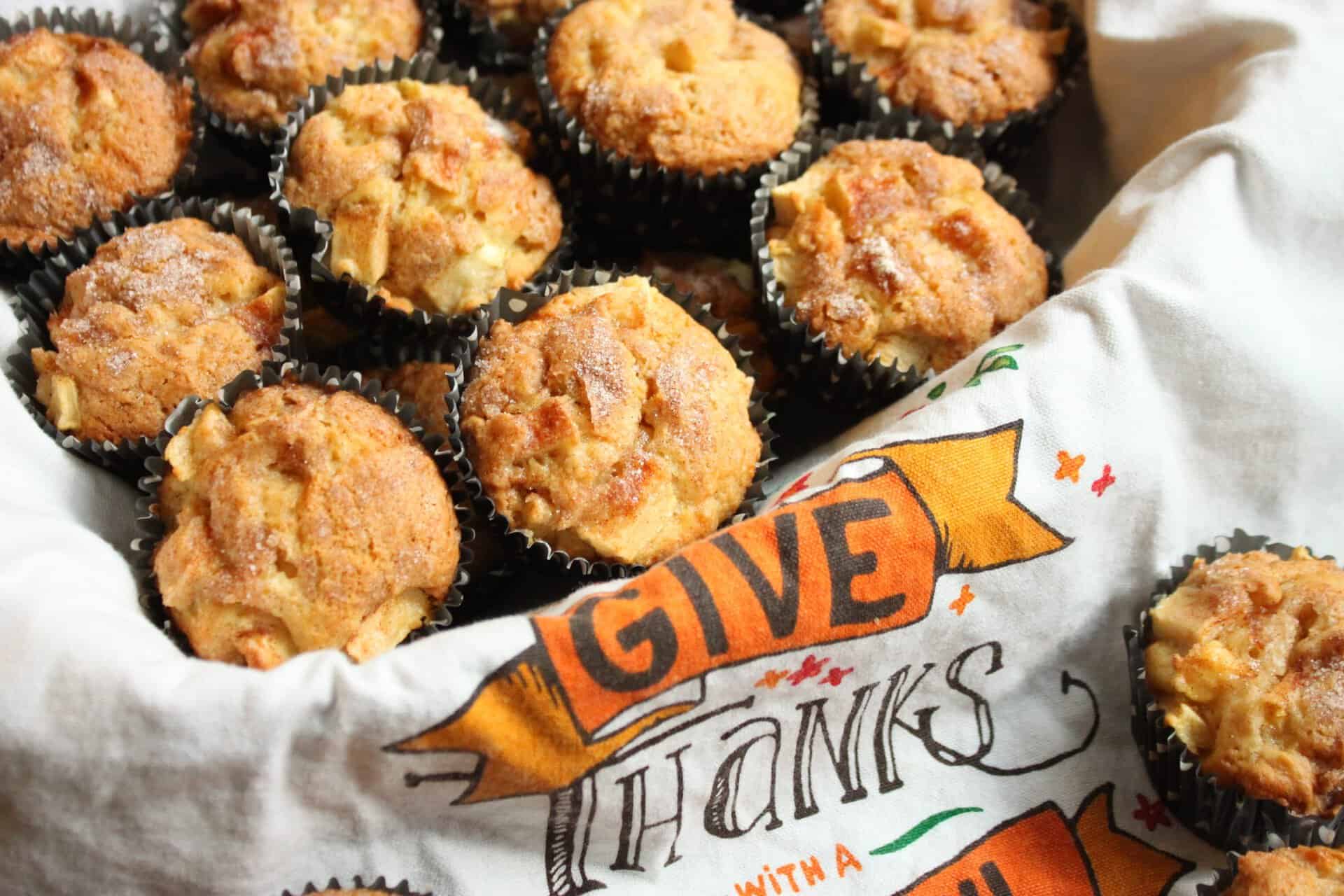 Cinnamon Apple Bread and Muffins: The Ultimate Fall Comfort Food 7 Close-up shot of golden cinnamon apple muffins nestled in black liners, topped with sugar, on a white napkin with an orange 'Give Thanks' banner.