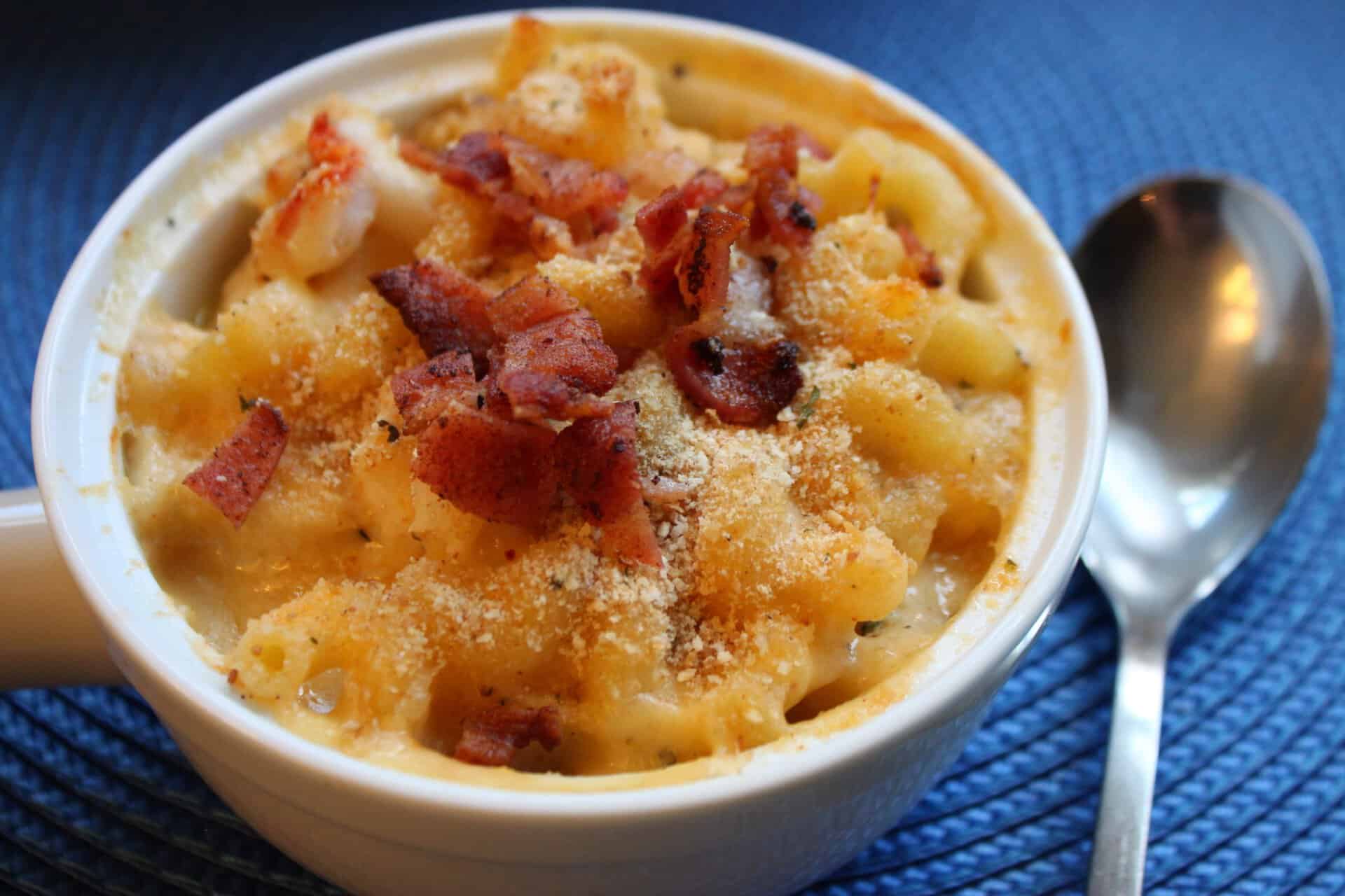 Individual ramekin of creamy lobster mac and cheese with a crispy, golden breadcrumb topping and crumbled bacon on a blue napkin.