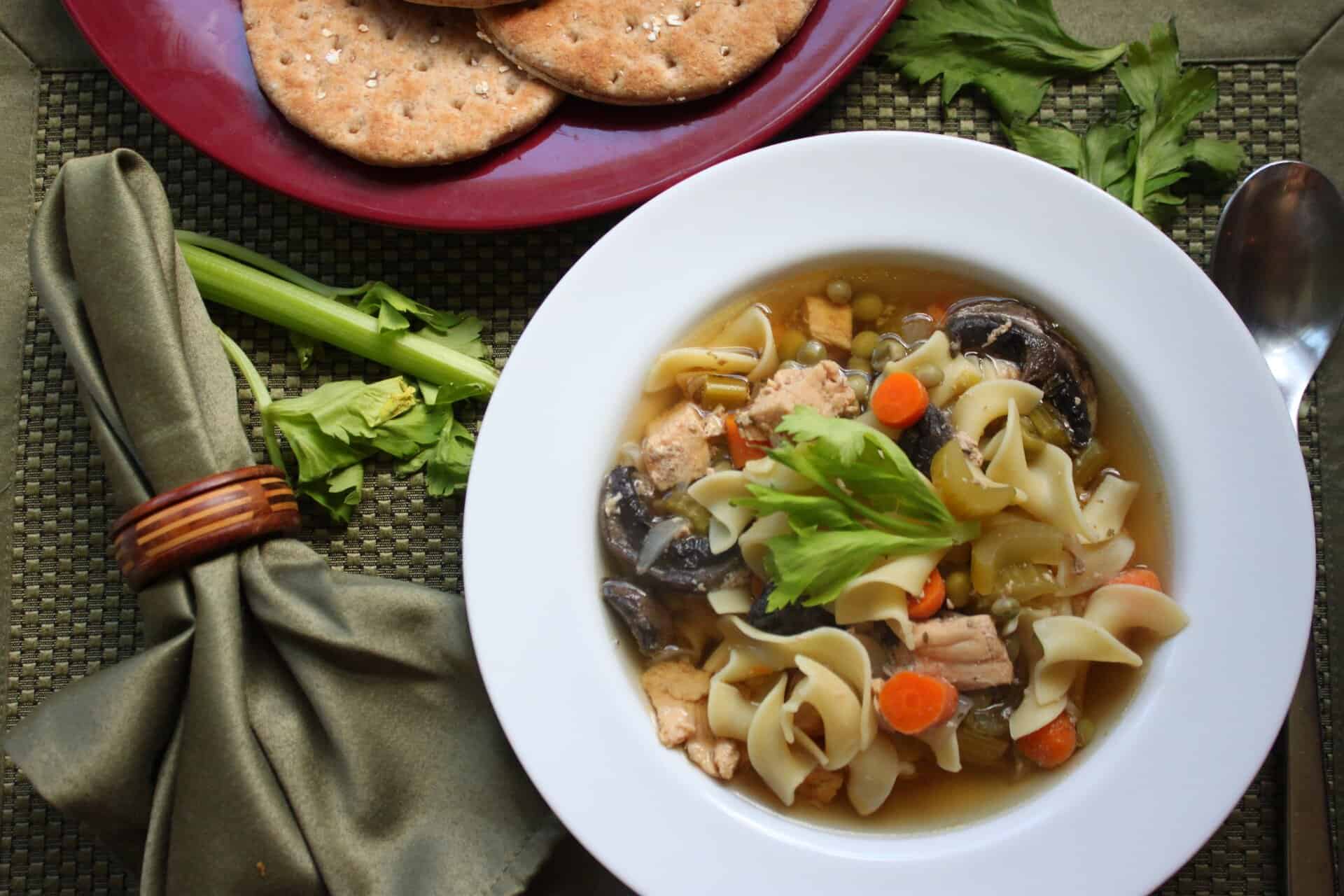Warm bowl of crockpot chicken noodle soup with carrots, mushrooms, and noodles, garnished with celery, next to crackers on a red plate.