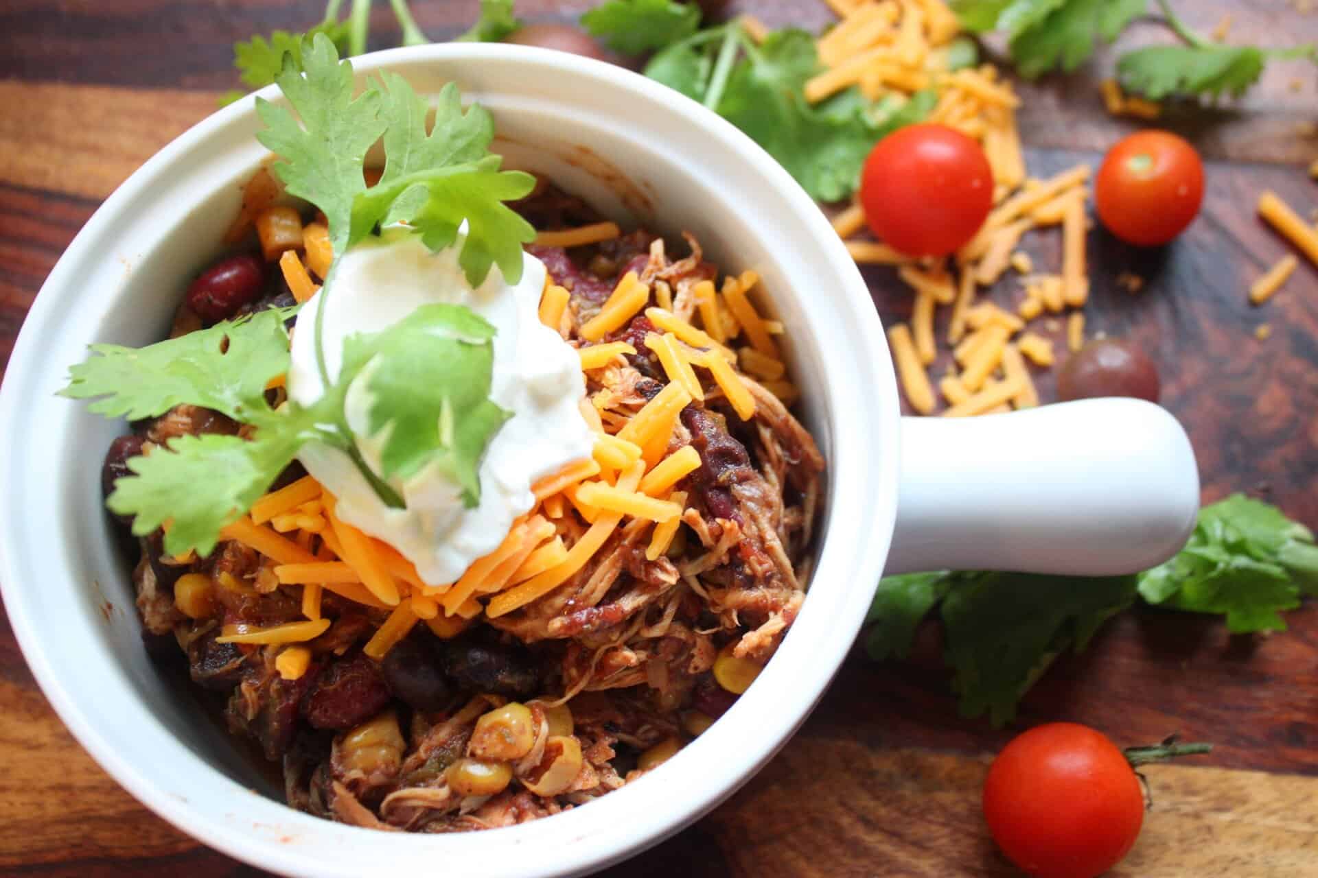How to Make Mouthwatering Crockpot Chicken Taco Chili 8 Close-up of Crockpot chicken taco chili in a white bowl with cheddar, sour cream, and cilantro, surrounded by tomatoes and shredded cheese.