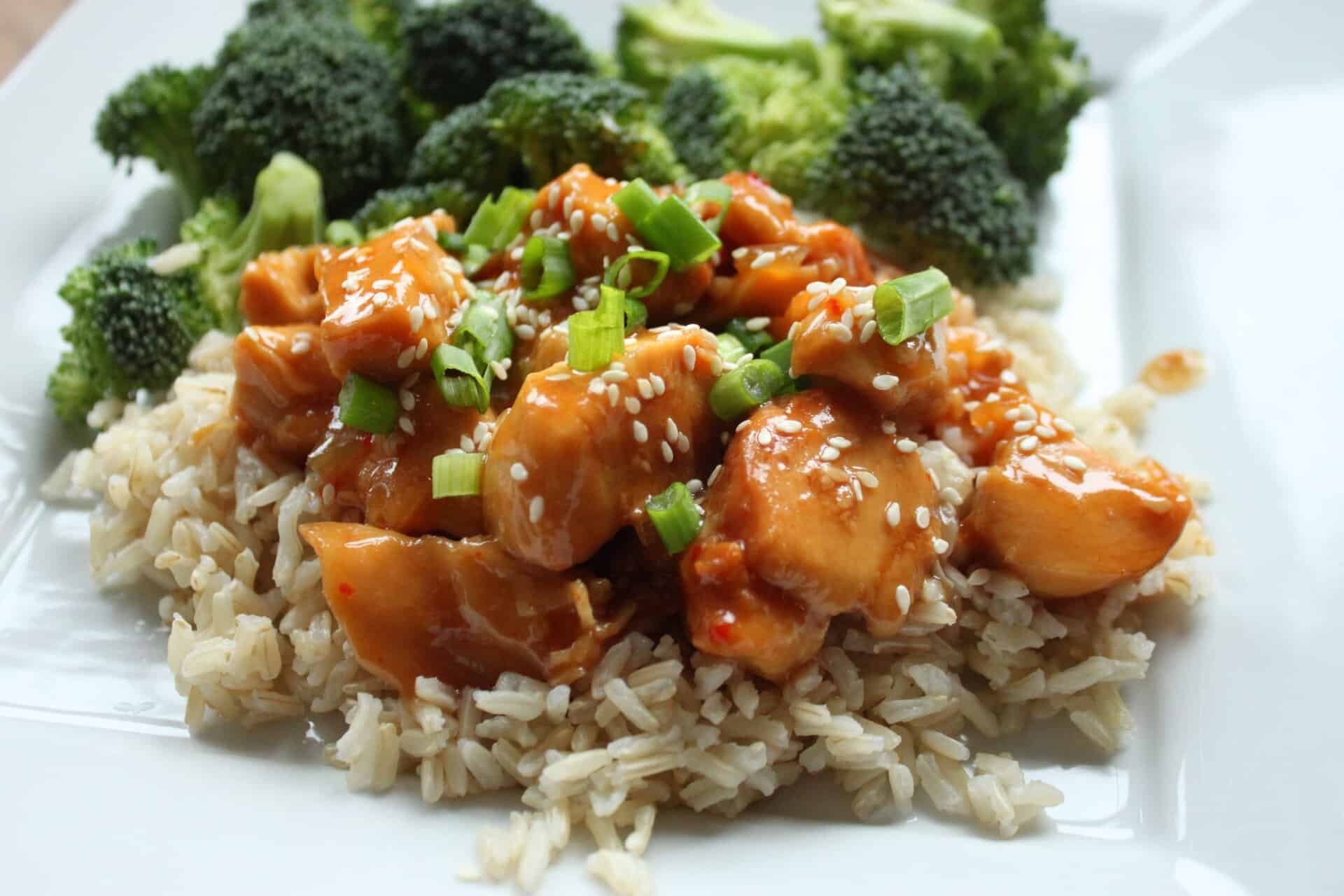 A hero shot of Easy Crockpot Sesame Chicken on a plate of rice, topped with sesame seeds and green onions, served with broccoli.