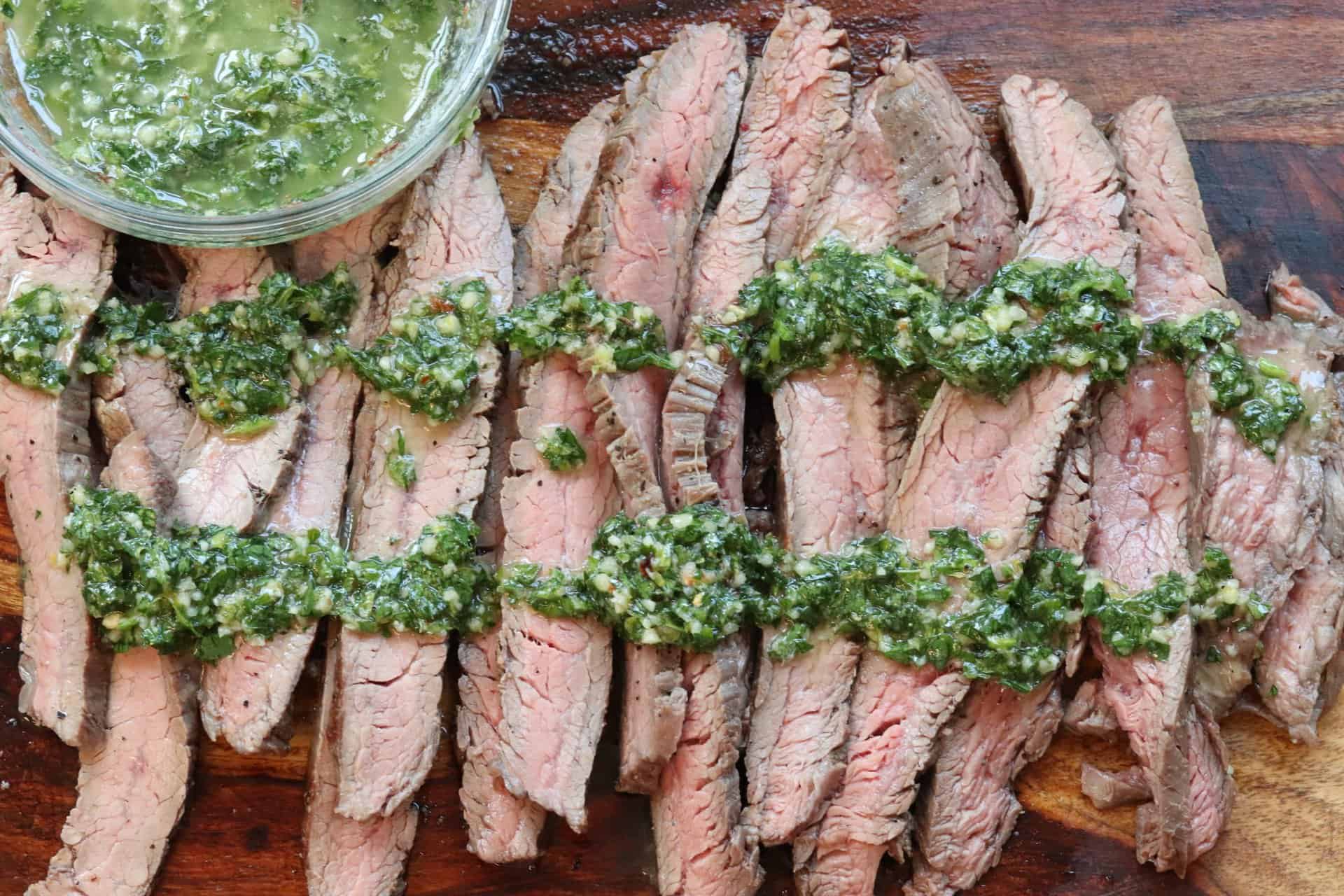 Grilled flank steak with chimichurri sauce served in thin slices, arranged in a row with bright green herb sauce on top and on the side.