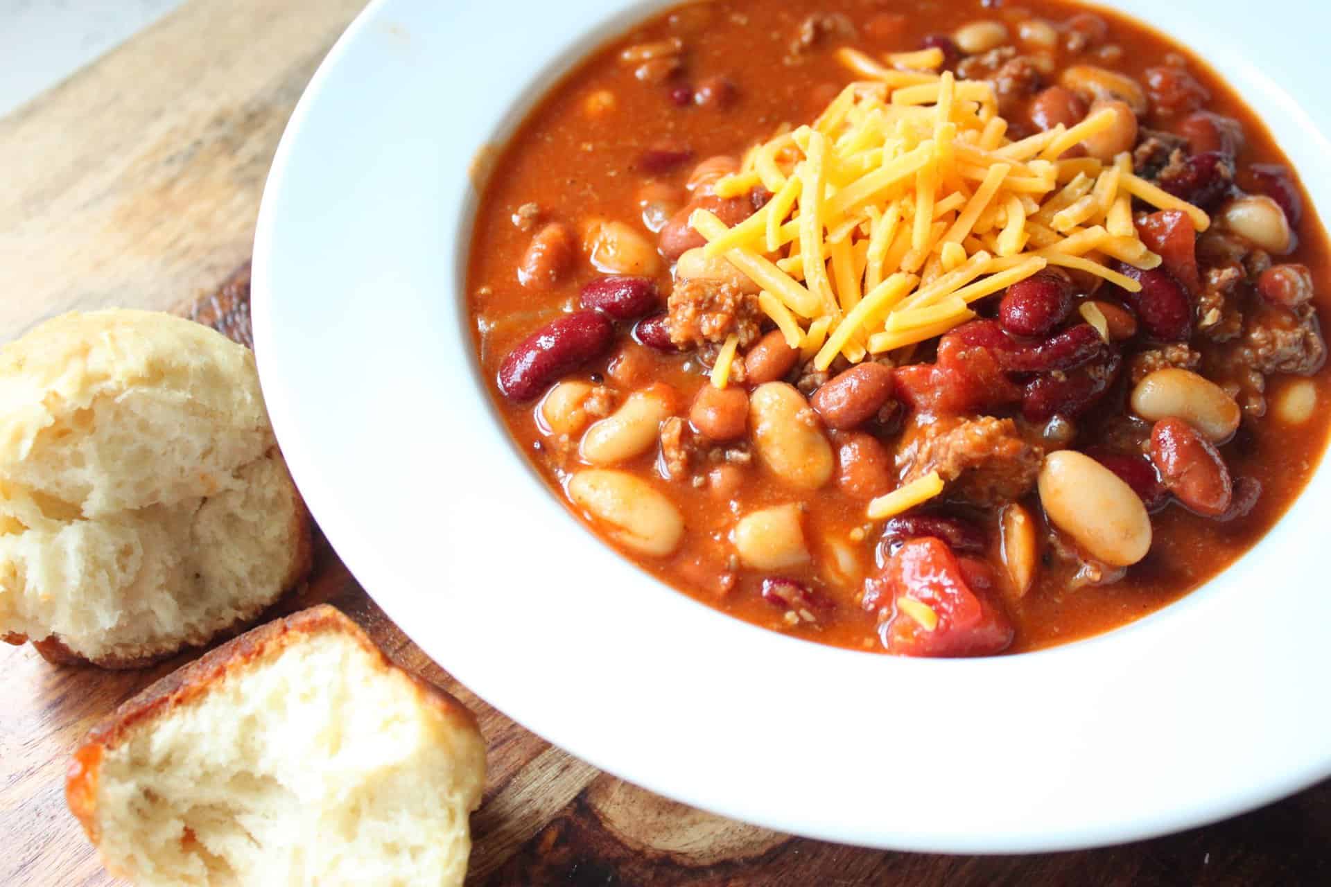 A bowl of hearty ground beef chili topped with shredded cheddar cheese, placed beside a slice of cornbread.