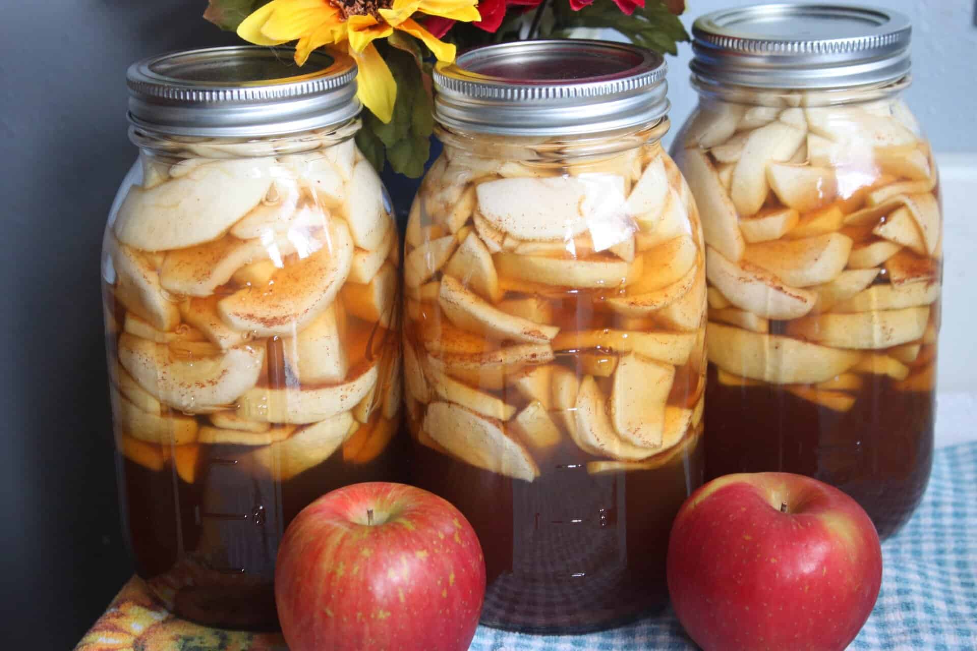 Three large mason jars filled with Homemade Canned Apple Pie Filling, surrounded by fresh apples and sunflowers for a fall-themed look.