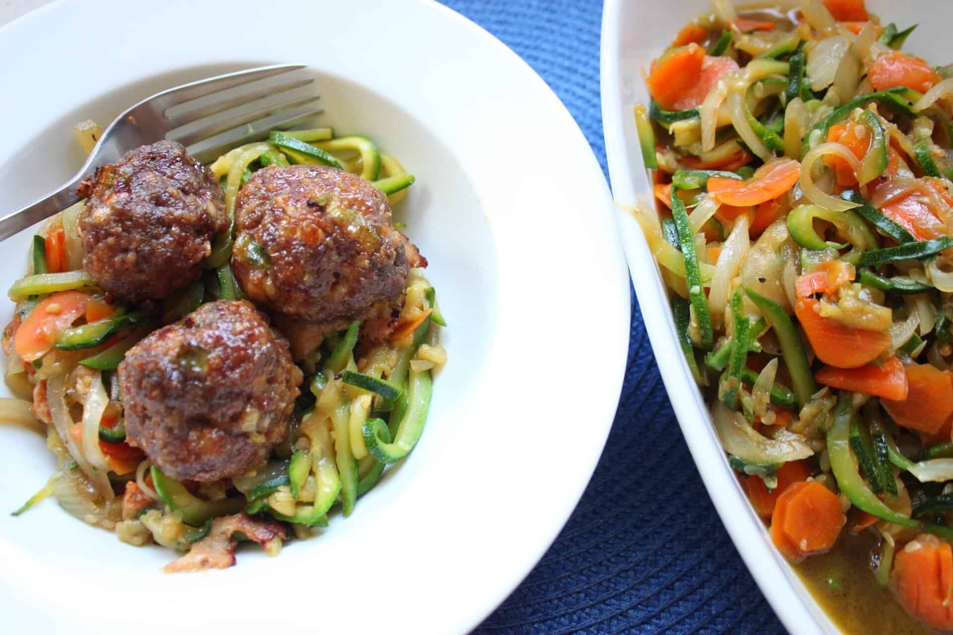Three Korean meatballs with stir-fry zucchini noodles in a white bowl, with a fork and extra veggies on the side.