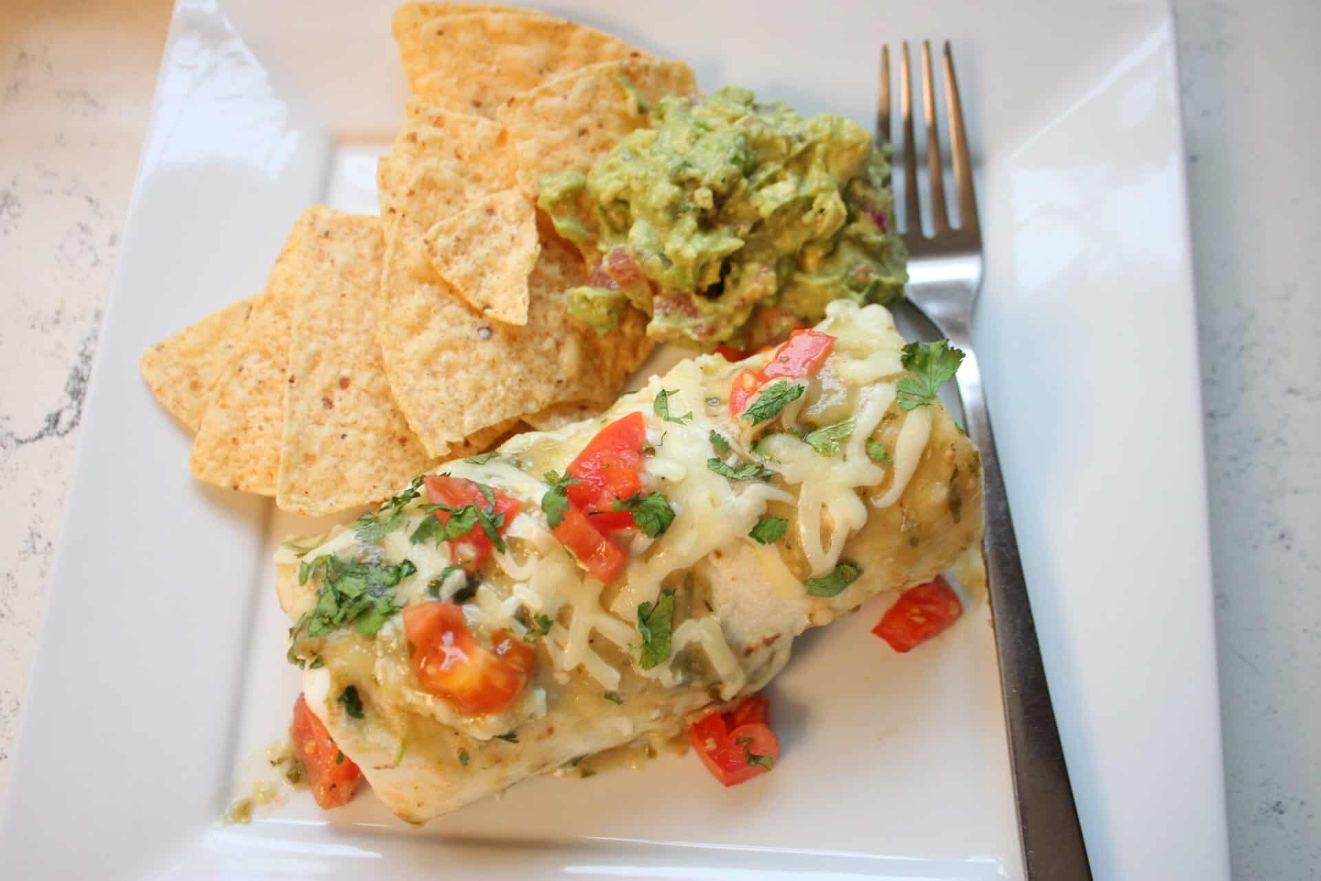 Plate of salsa verde chicken enchiladas topped with melted cheese and diced tomato, served with chips and guacamole on the side.