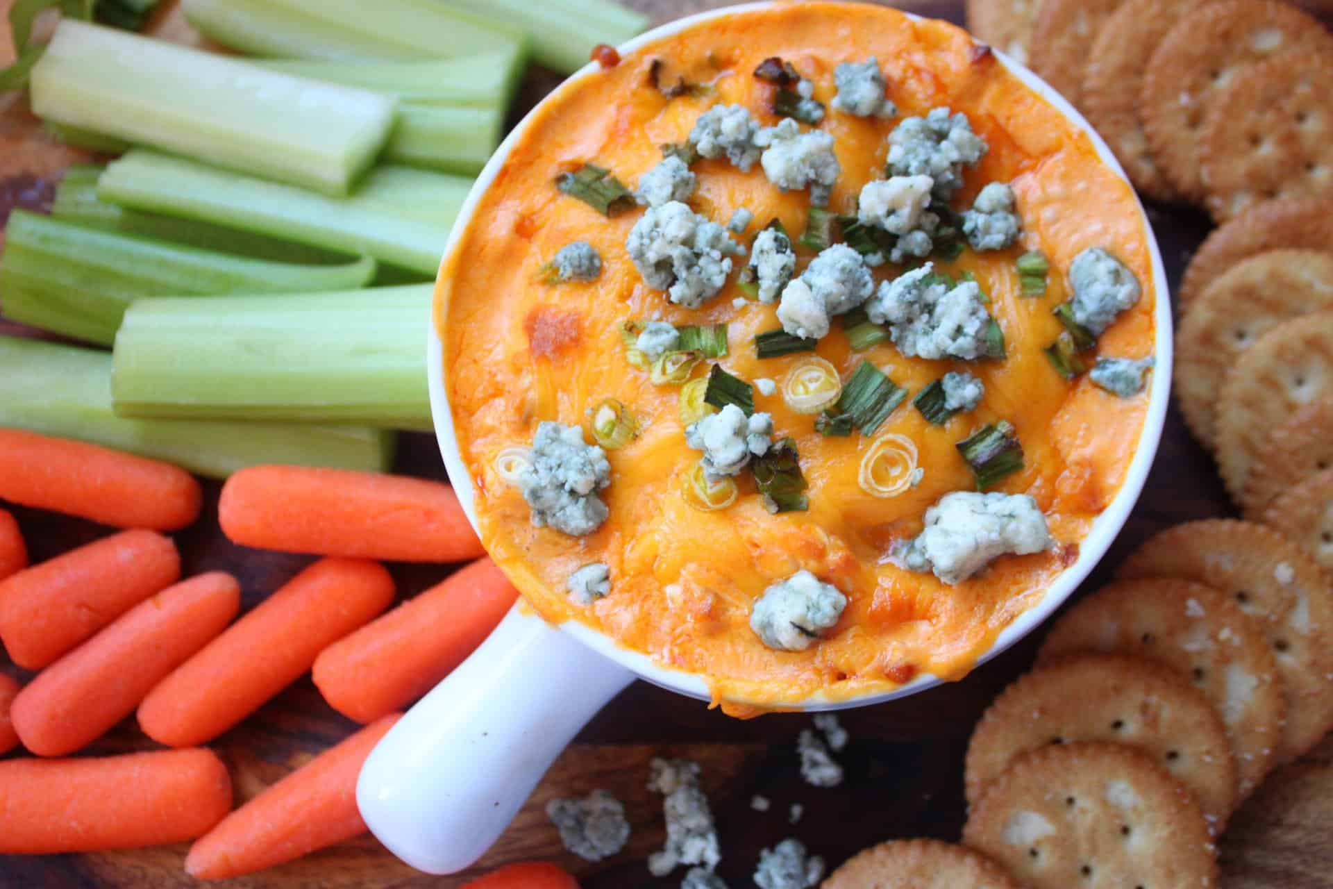Overhead view of spicy buffalo chicken dip in a white dish, topped with bubbly cheddar, chopped scallions, and crumbled blue cheese.