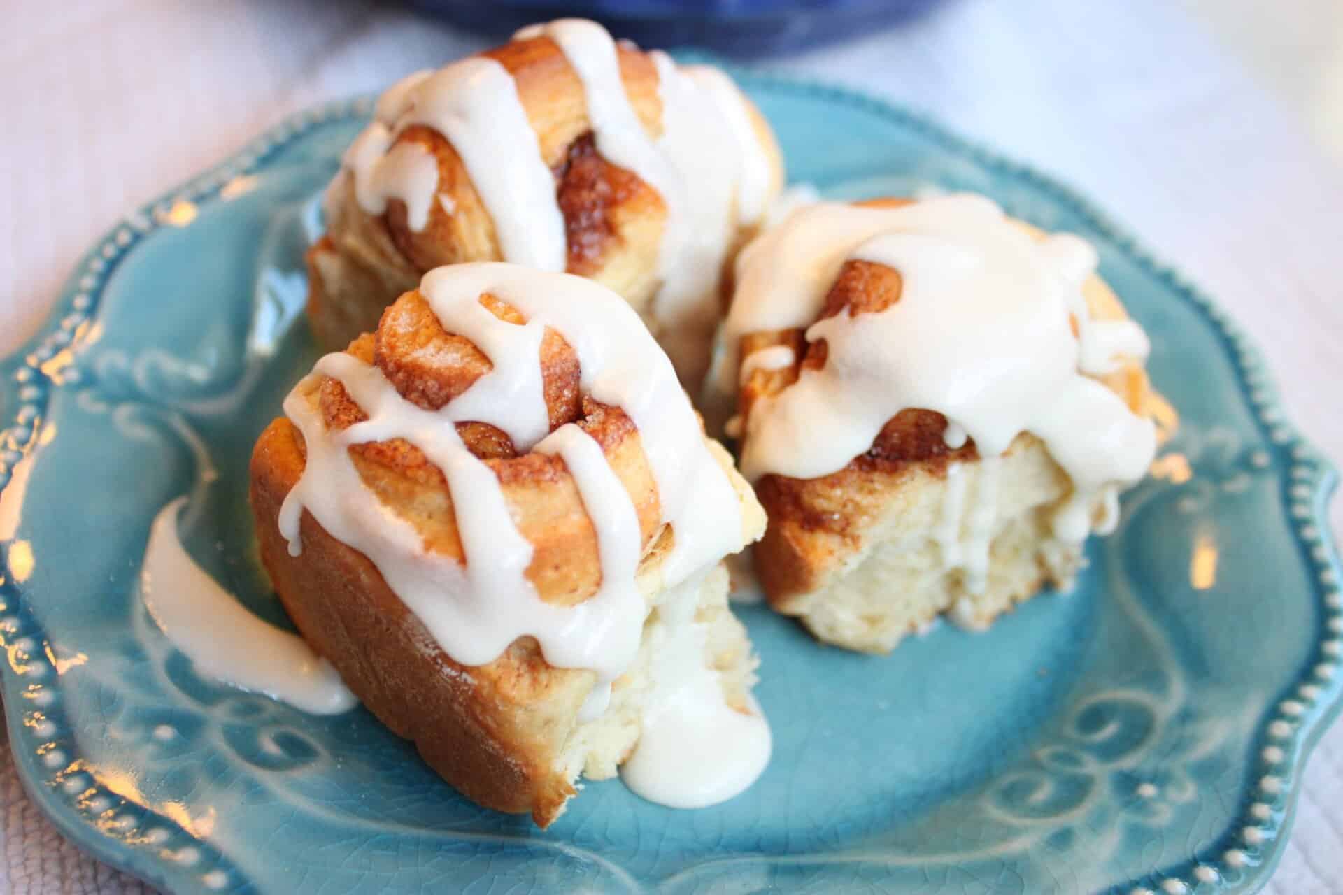 Three iced homemade cinnamon rolls on a blue plate, each roll topped with thick, creamy frosting and full cinnamon swirl.