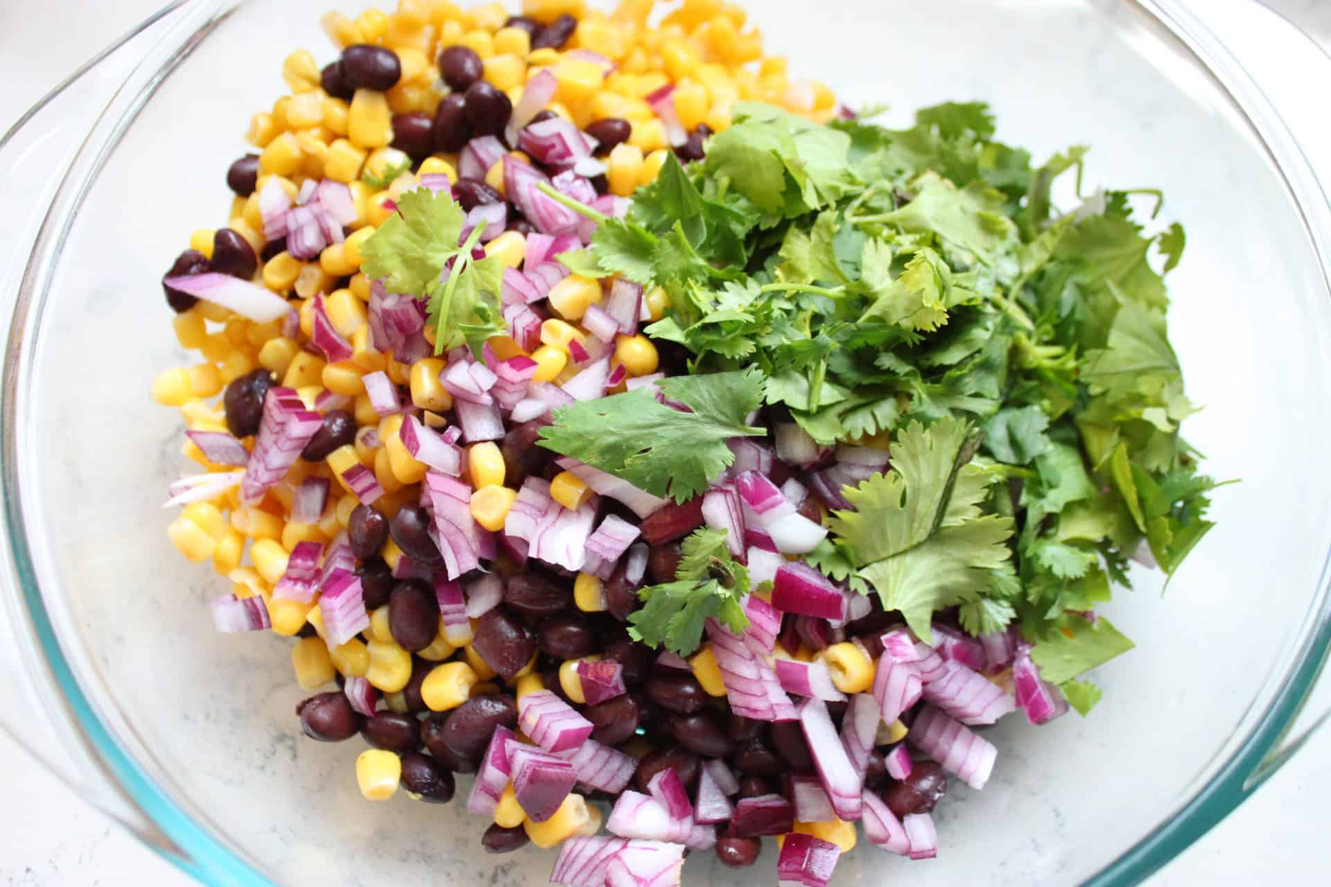 Chopped red onion, corn, black beans, and fresh cilantro in a bowl before mixing, the starting ingredients for black bean and corn salad.