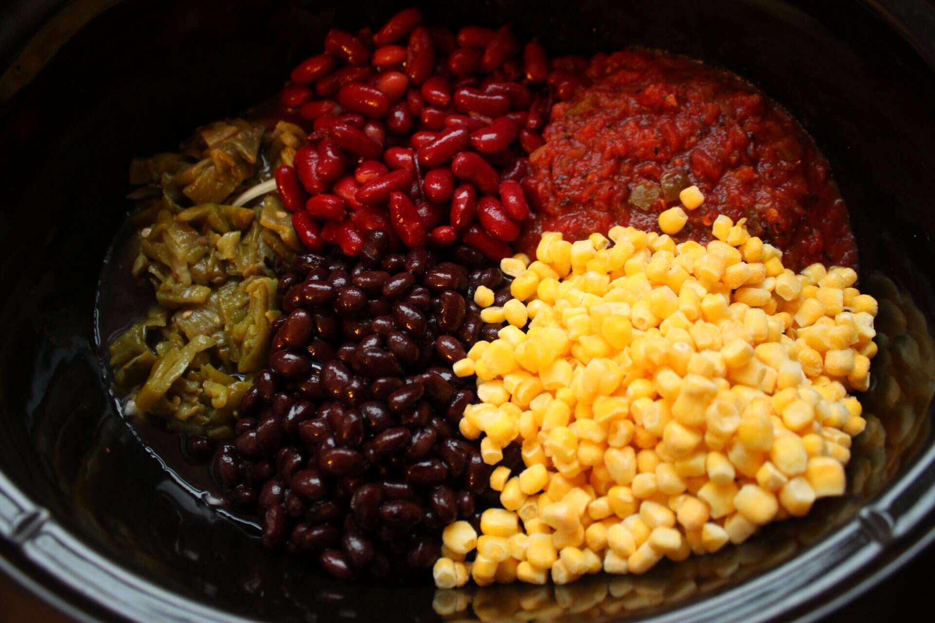 Unmixed Crockpot chicken taco chili ingredients including beans, corn, and salsa layered in a slow cooker before combining.