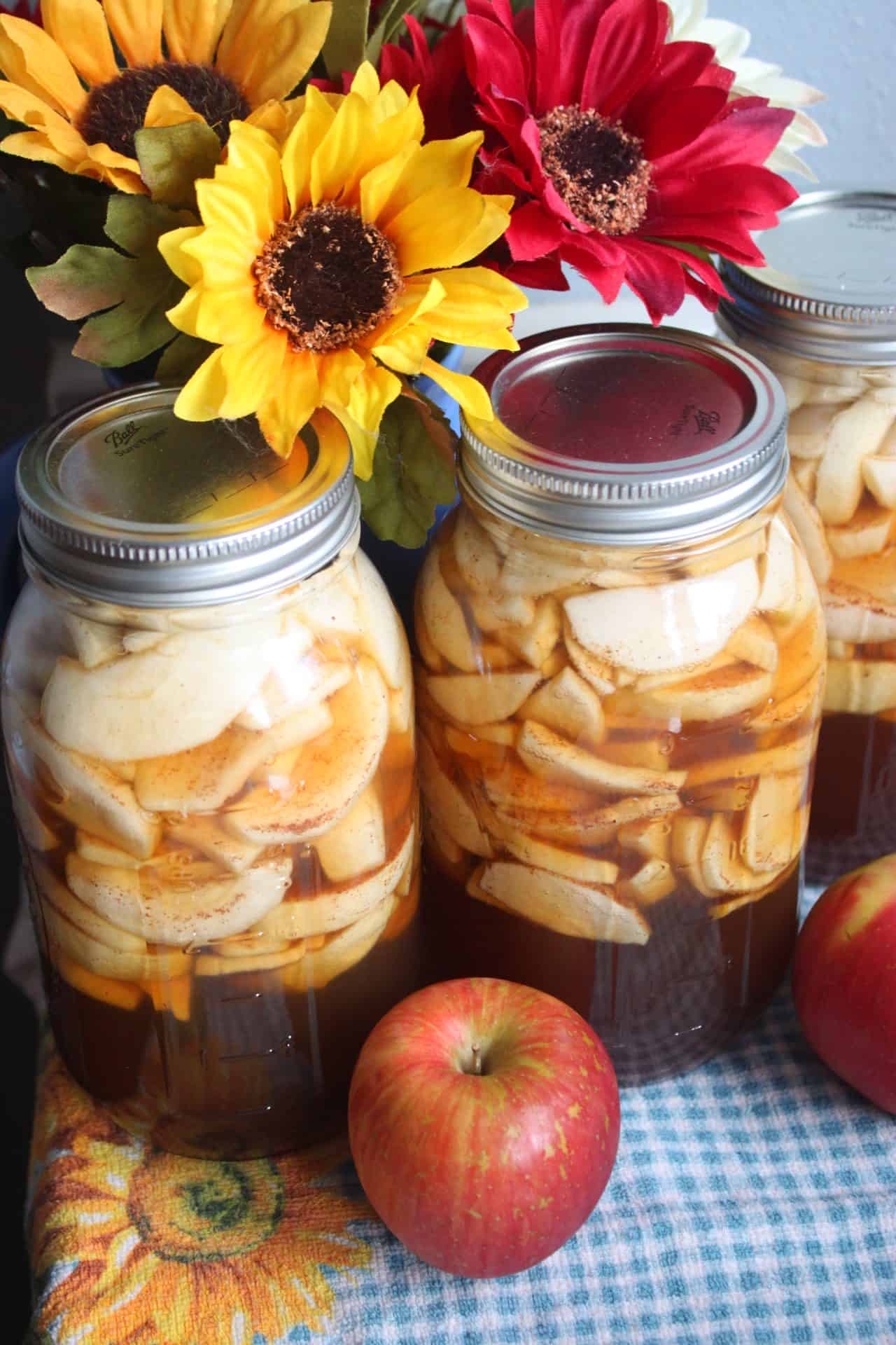 Close-up of two jars of Homemade Canned Apple Pie Filling with sliced apples and syrup, topped with silver lids and surrounded by fresh sunflowers.
