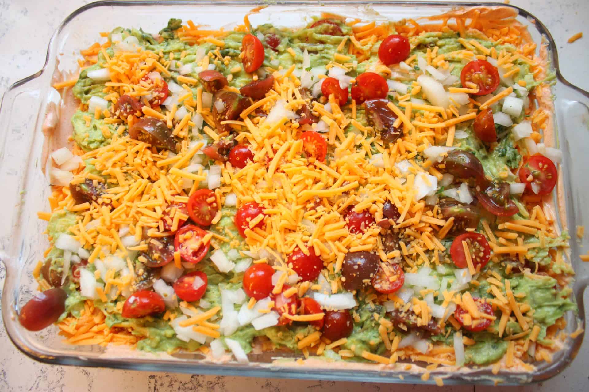 Crowd-Pleasing 7-Layer Mexican Bean Dip for Dinner or Parties 2 Top-down view of a glass dish filled with colorful layers of 7 Layer Mexican Bean Dip, featuring shredded cheese, tomatoes, and guacamole.