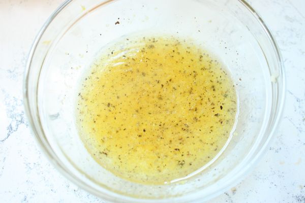 Glass bowl of freshly whisked lemon dressing with visible herbs, garlic, and lemon zest for adding to lemon chicken orzo salad.