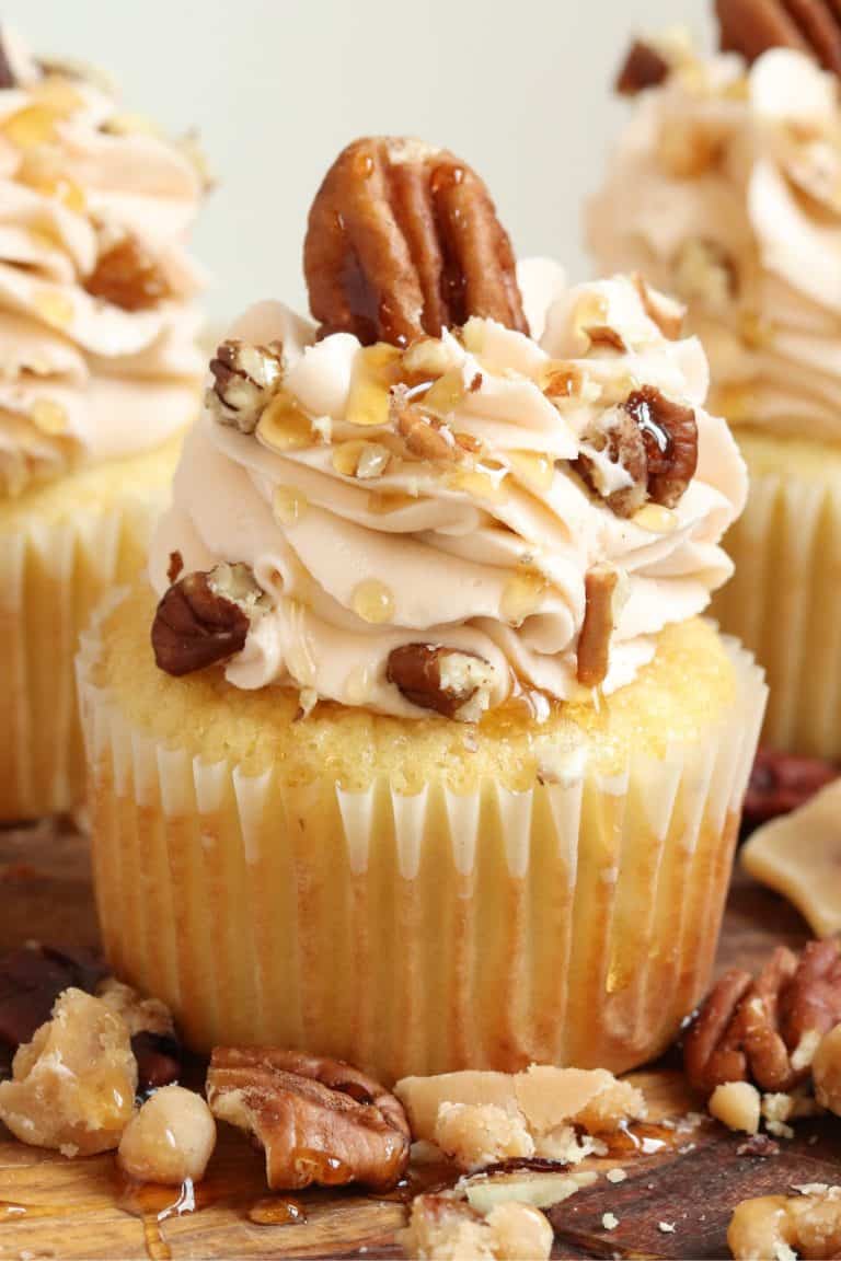 A cupcake with swirled buttercream, topped with a large pecan and syrup drizzle, adorned with crushed pecans, creating a rustic pecan theme.