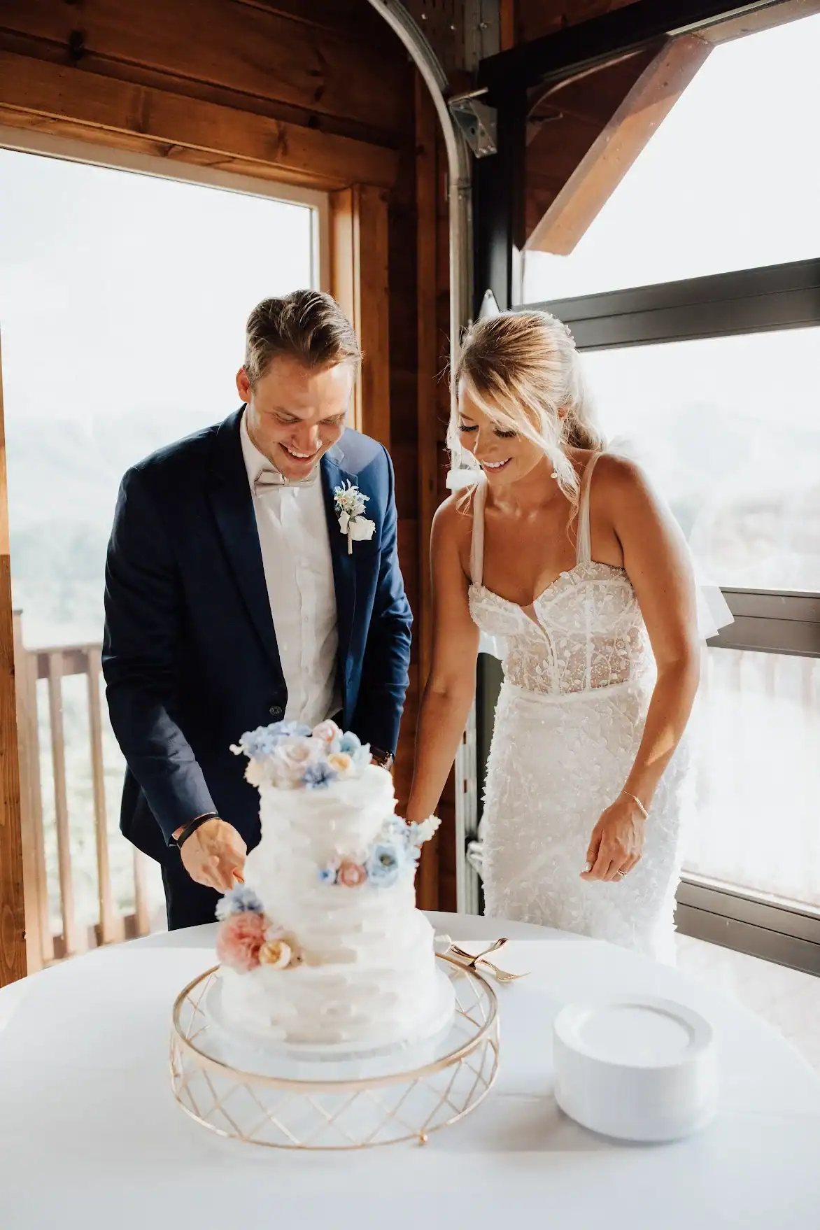 White tiered wedding cake adorned with pastel-colored flowers, with a bride and groom about to cut the cake together.