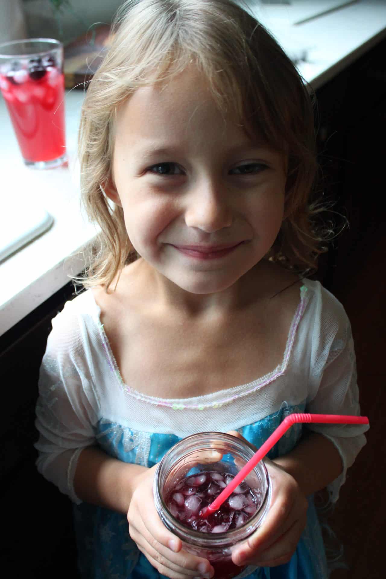 Smiling child with a glass of Starbucks Iced Passion Tea Lemonade on a windowsill, enjoying a refreshing summer drink.