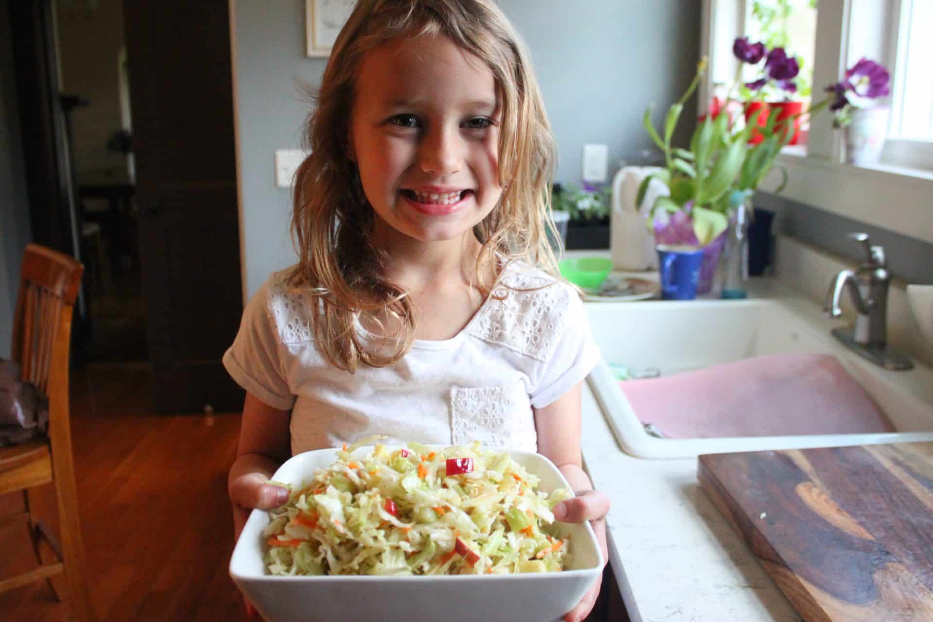This Crisp Sweet Apple Celery Coleslaw is A Southern Favorite 6 A young girl in a white shirt holding a bowl of Sweet Apple Celery Coleslaw, smiling proudly in a kitchen setting.