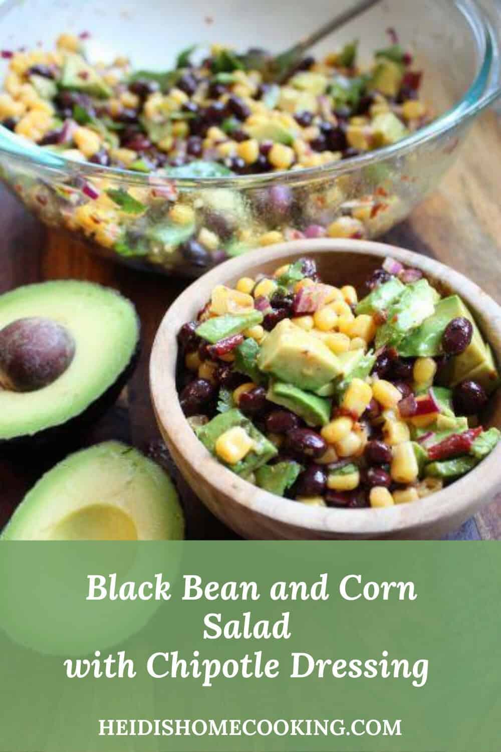Large glass bowl and small wooden bowl of black bean and corn salad with chipotle dressing, surrounded by fresh avocado halves on a wood surface.
