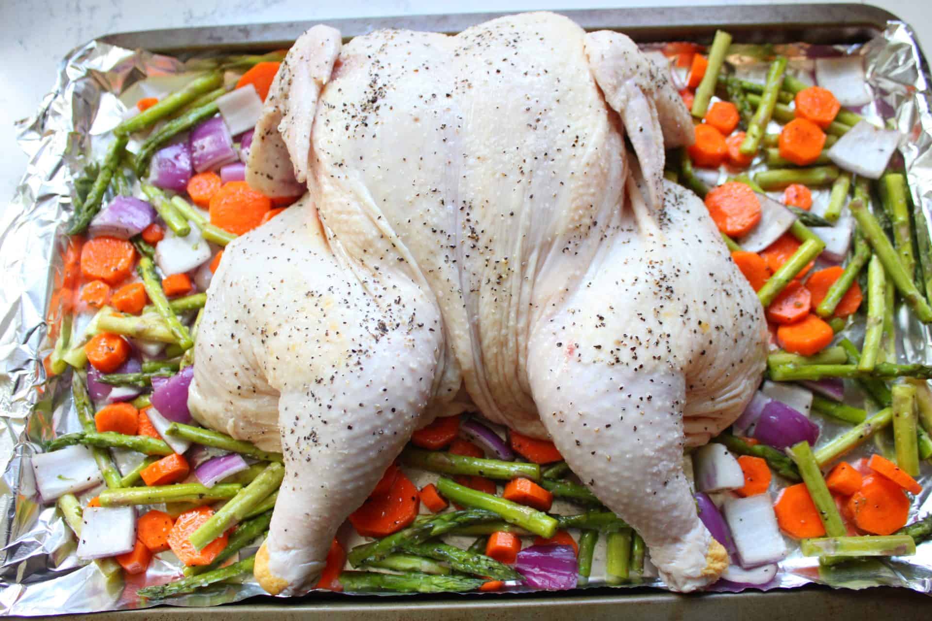 Uncooked spatchcock chicken placed on a bed of chopped vegetables on a foil-lined sheet pan before roasting.