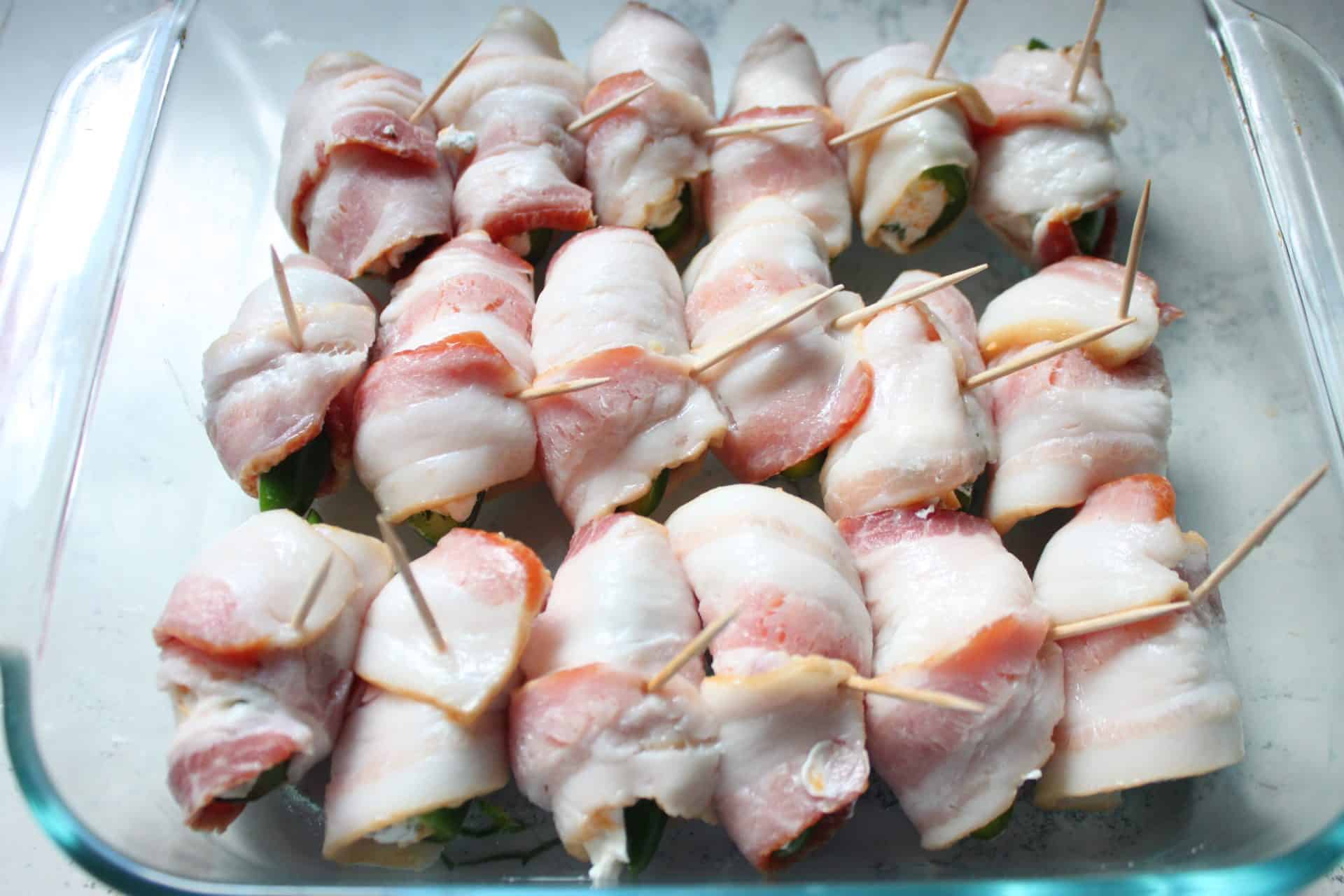 Crispy and Creamy Easy Bacon Wrapped Jalapeño Poppers Recipe 6 Rows of bacon wrapped jalapeño poppers secured with toothpicks on a tray, ready to be cooked until crispy and delicious.