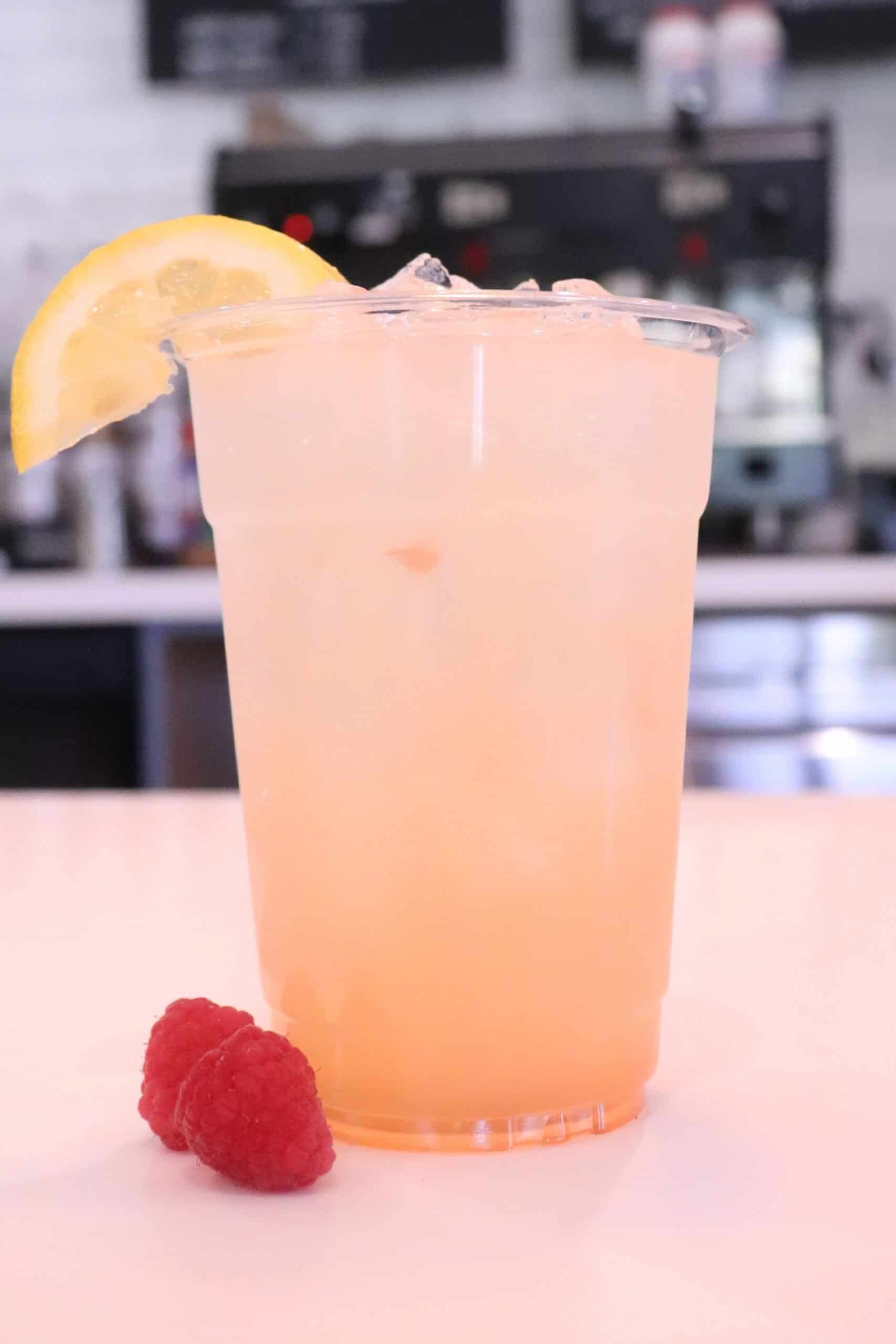 Chilled raspberry lemonade with ice, a lemon wedge garnish on the rim, and two raspberries placed next to the cup in the foreground.