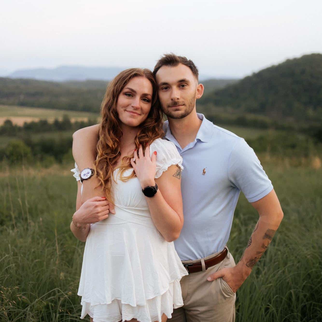 A couple stands close together, with the woman in a short white dress and the man in a light blue collared shirt with tan pants, arms wrapped around each other.