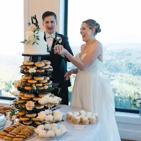 Tall tower of assorted wedding desserts including layered cakes, pastries, and sweets, with a bride and groom standing beside the display.