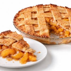 Peach pie slice with juicy filling spilling onto a white plate and the whole lattice pie with one slice missing behind it.