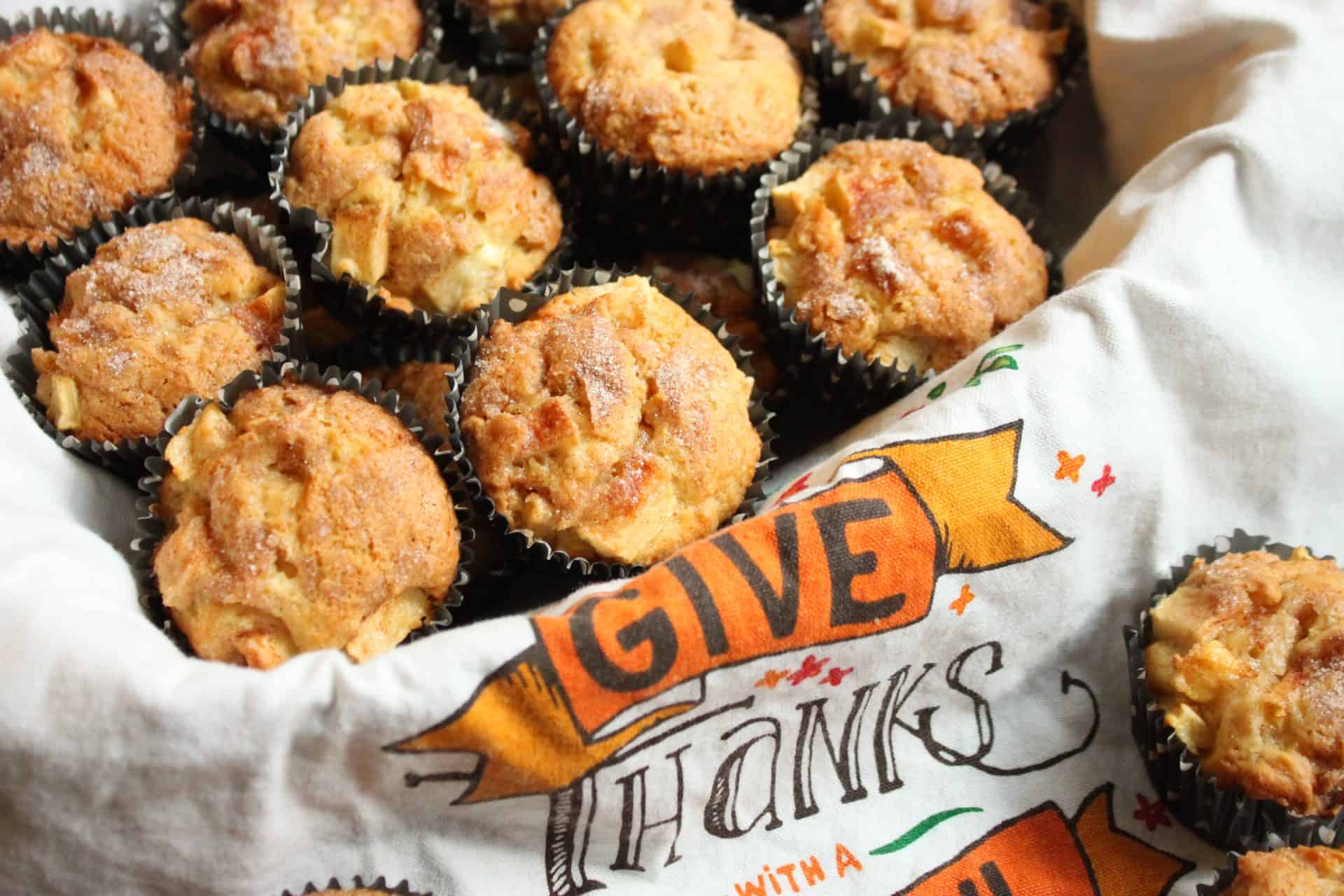 A serving dish filled with baked cinnamon apple muffins, arranged on a white napkin with an orange 'Give Thanks' banner for a festive touch.