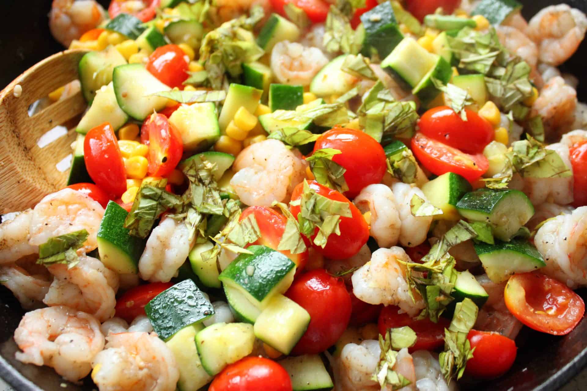 Stirring Together Shrimp, Corn, Cherry Tomatoes, Zucchini and Basil in a Crockpot with a Wooden Spoon