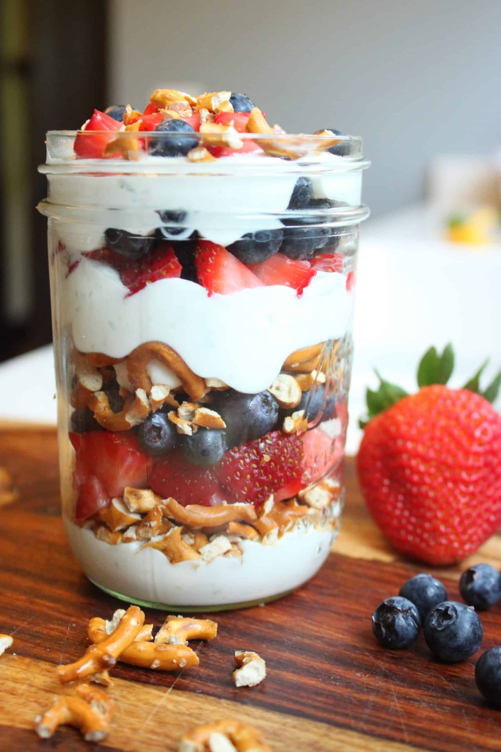 Irresistible Patriotic Lime Yogurt Parfaits for Fourth of July Fun 7 Side View of Patriotic Lime Yogurt Parfait with Blueberries, Strawberries, and Crushed Prezels spread around.